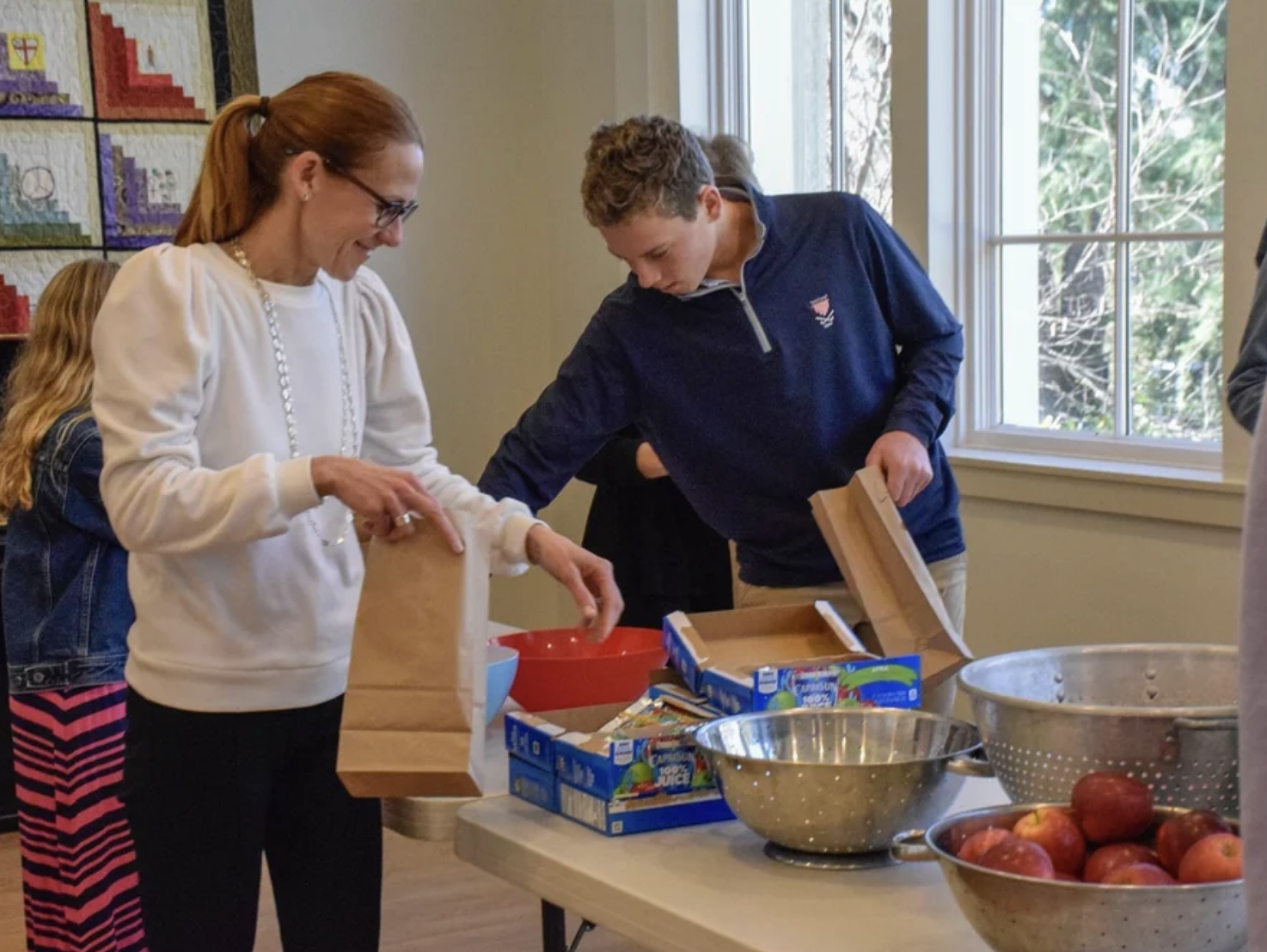 A woman with red hair and glasses, wearing a white sweater and a pearl necklace, is packing items into a paper bag at a table. A boy in a navy jacket is opening a box of fruit snacks on the table. The table also has bowls of apples and snacks, and people are in the background near a window.
