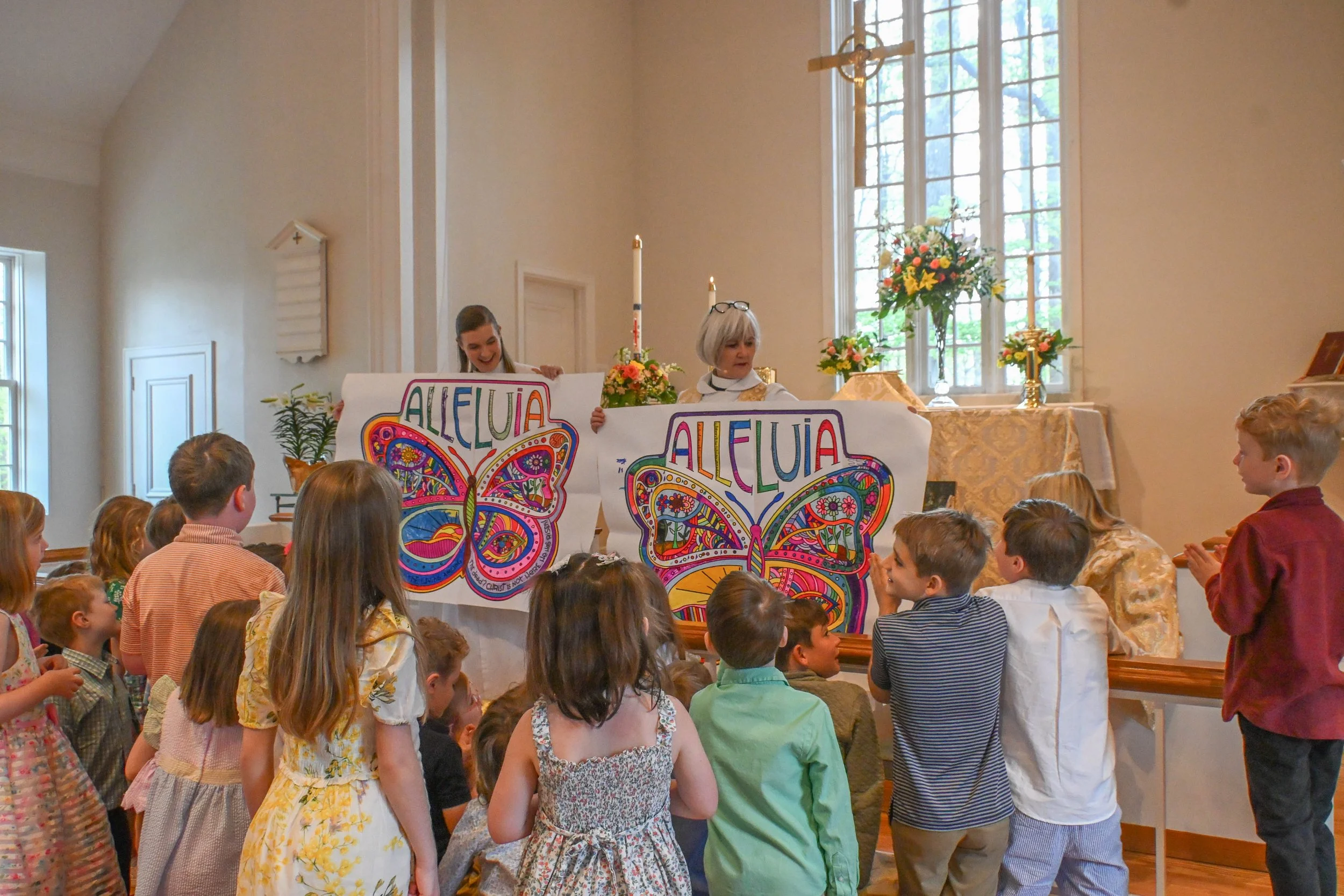 Children gathered inside a church during a celebration, with two women holding large colorful butterfly posters with the word 'ALLELUIA' on them. The church has large windows, floral arrangements, and a wooden cross hanging from the ceiling.