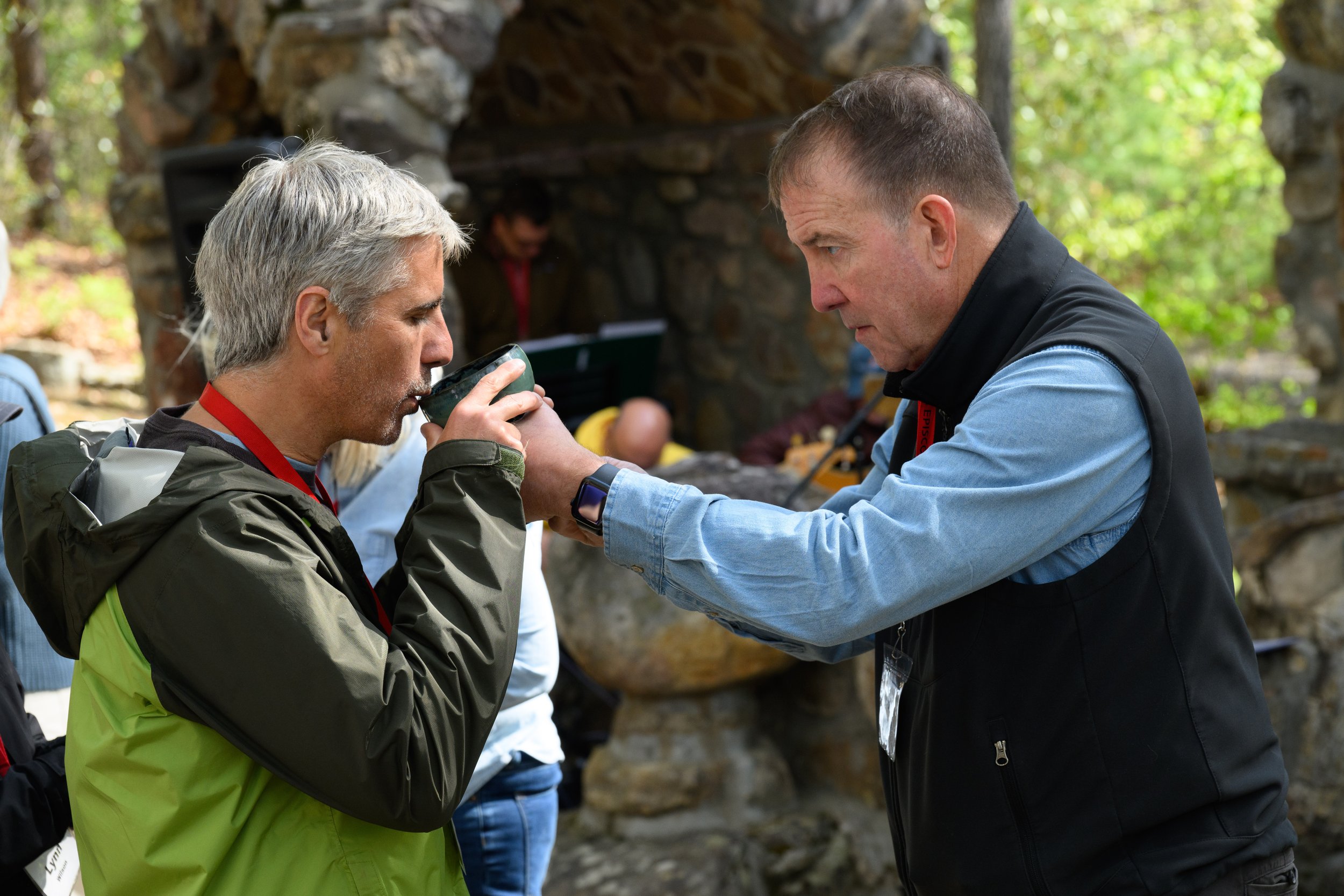 Two men outdoors, one man is drinking from a cup, and the other is holding the cup to his lips. The scene appears to be in a wooded area with rocks and a stone structure in the background.