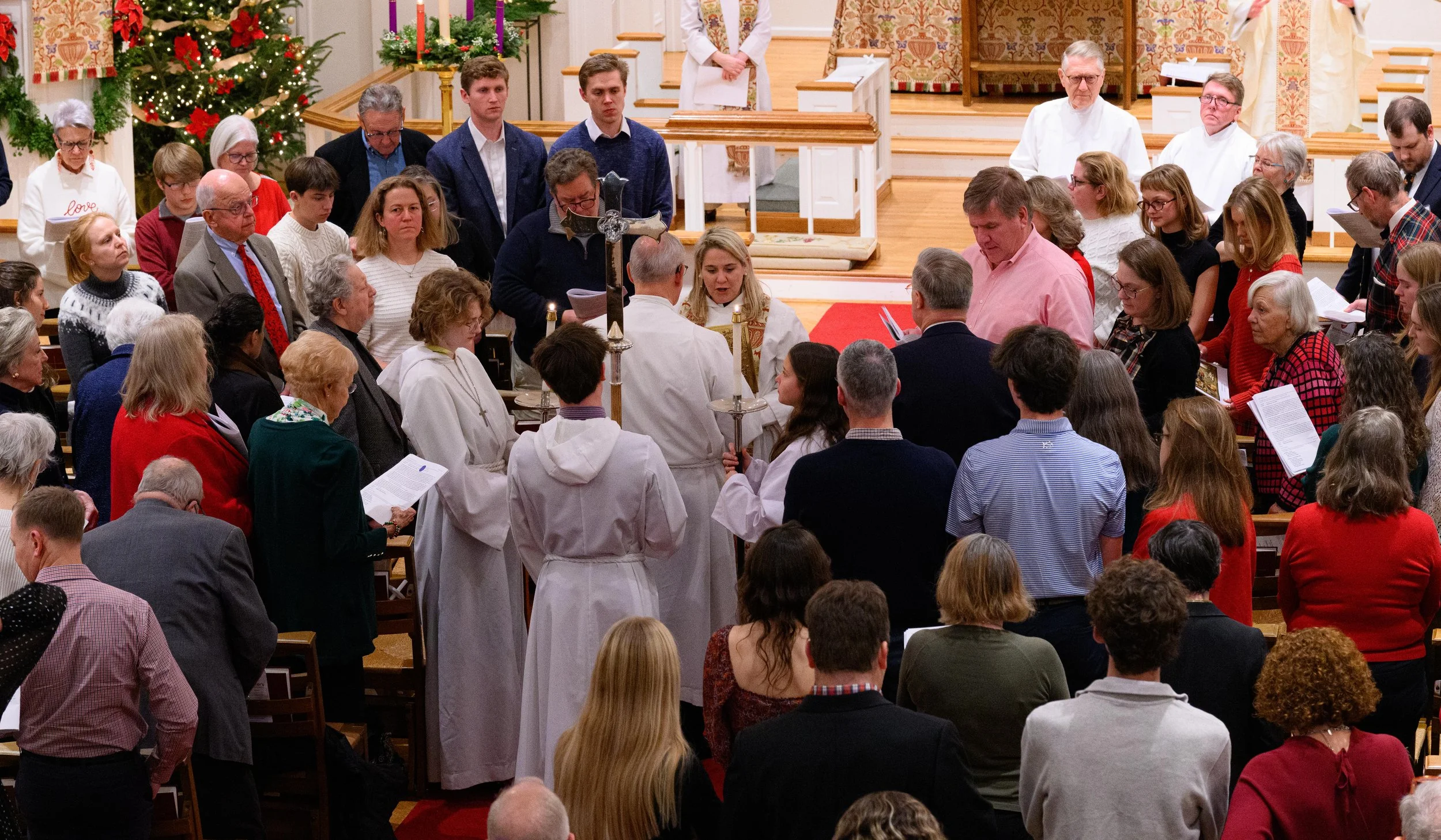 A church congregation gathered for a service with a Christmas theme, featuring a decorated Christmas tree with red ornaments and candles, people praying and singing, and clergy in white robes.