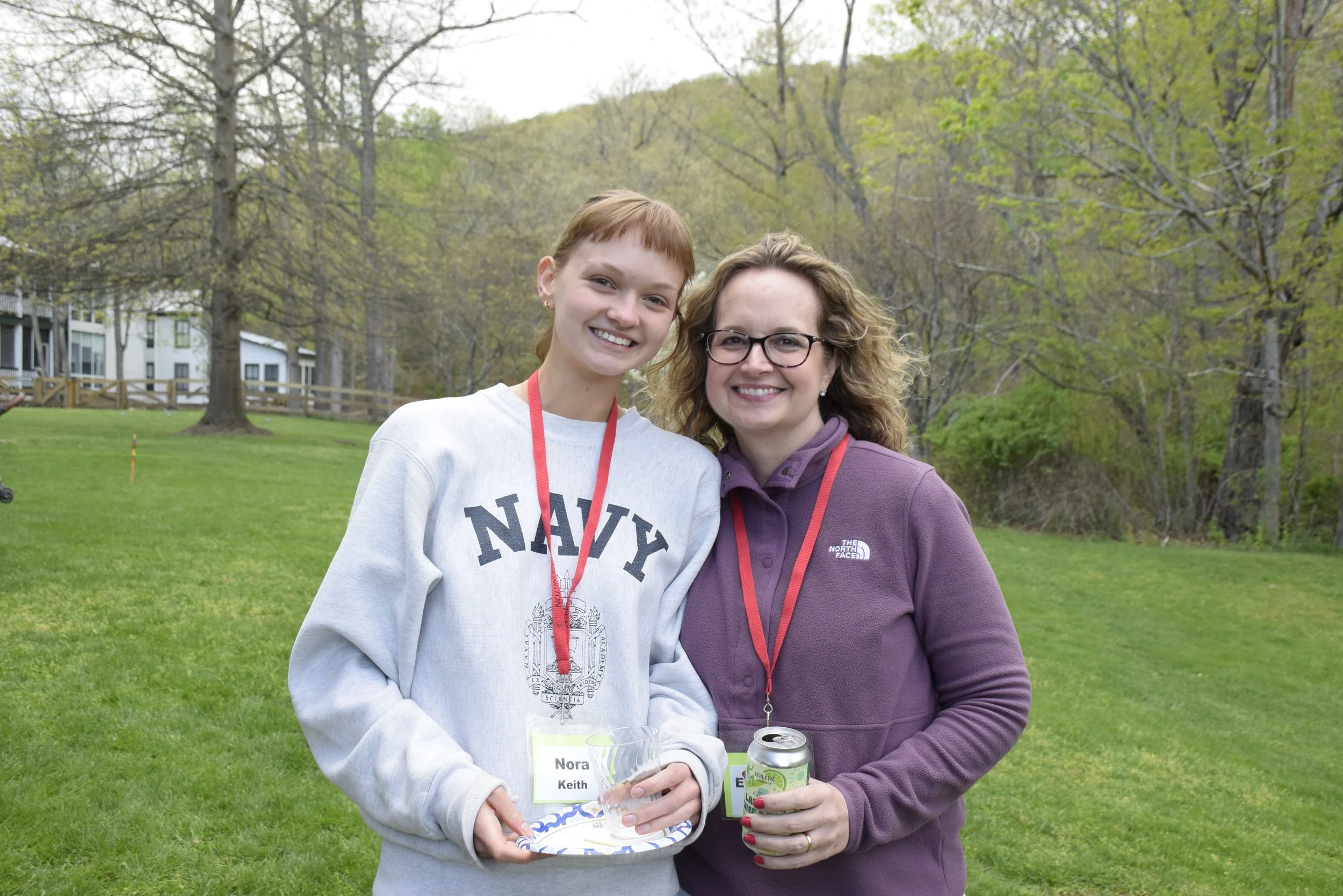 Two women standing outdoors on a grassy area, smiling at the camera. The woman on the left has short red hair, is wearing a gray sweatshirt with 'NAVY' on it, and has a red lanyard. The woman on the right has curly light brown hair, glasses, and is wearing a purple jacket with a red lanyard. They are holding drinks, one with a clear cup and the other with a can, and there are trees and houses in the background.