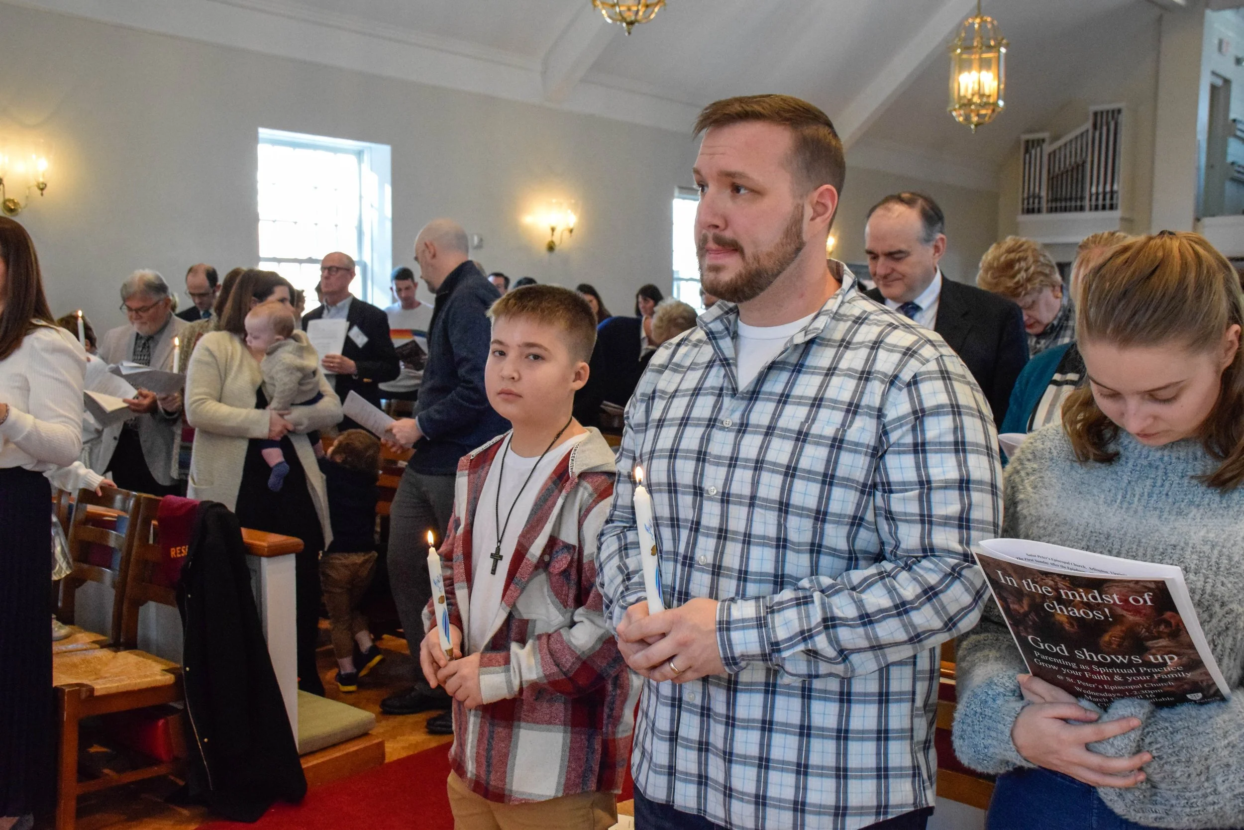 People standing inside a church during a religious service, holding candles, with some reading pamphlets or prayer books.