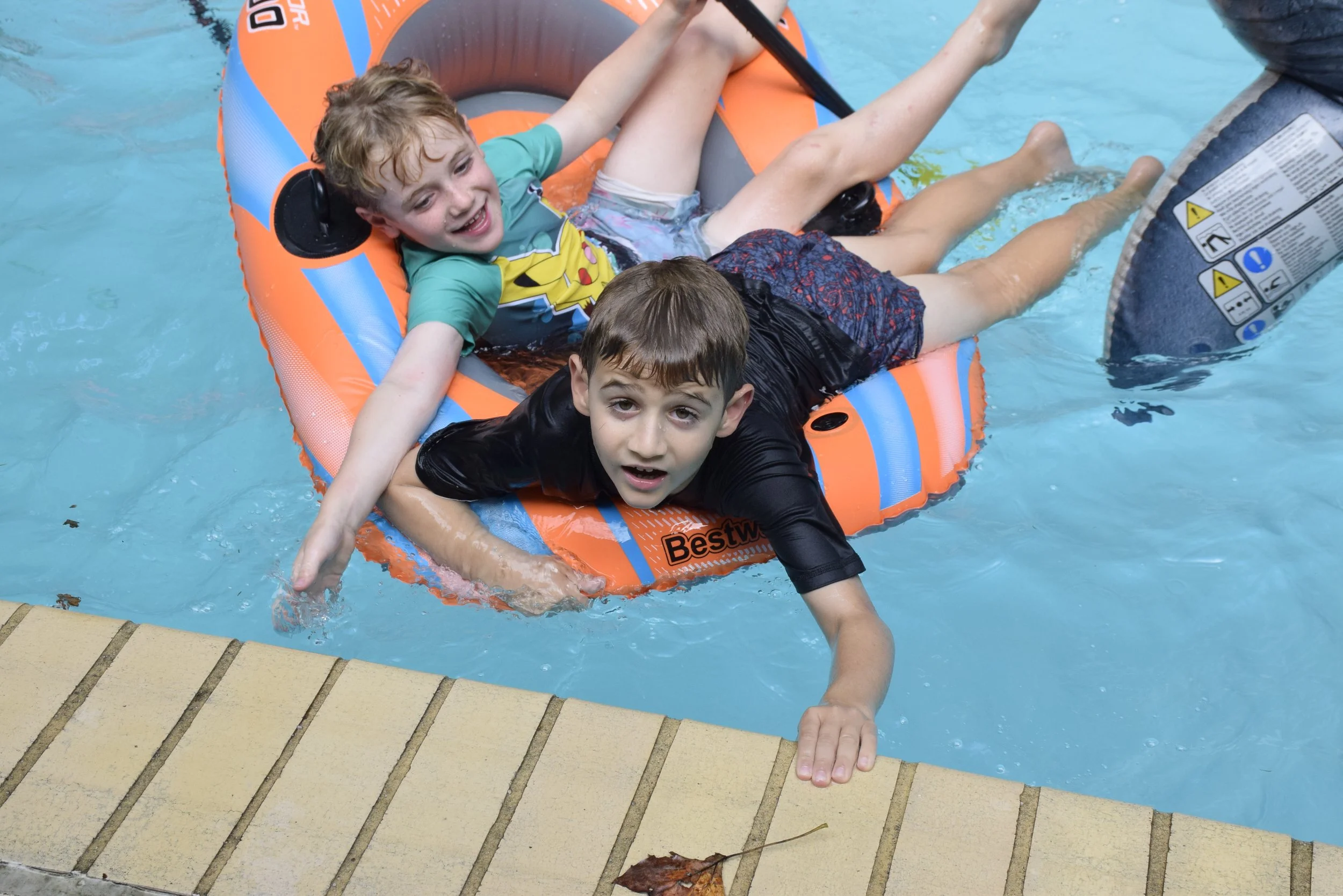 Two young boys in a swimming pool, one climbing out with a hand on the pool's edge, the other lying on an orange and blue inflatable raft in the water. The boy on the raft is smiling, and the other looks surprised or curious. An adult's leg is partially visible near the raft, and a leaf is on the pool's edge.