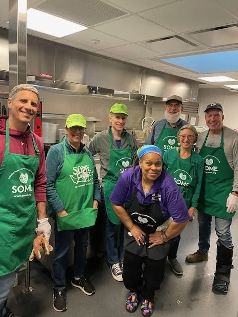 Group of five volunteers in a commercial kitchen, wearing green aprons and face masks, preparing food for a community meal program.