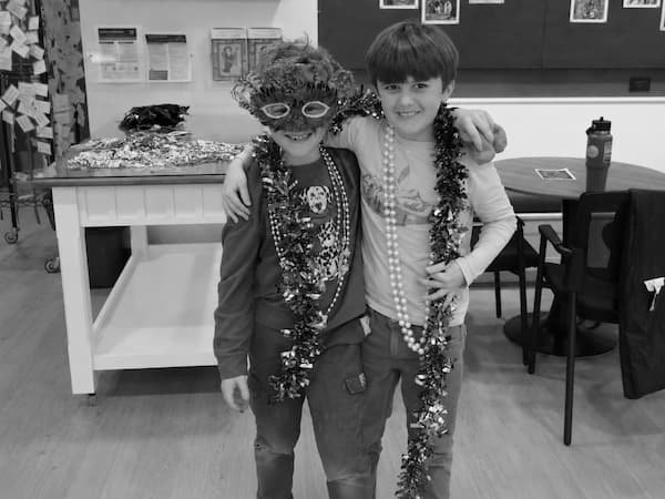 Two children dressed up with tinsel and accessories, smiling and posing together in a room with tables and chairs.