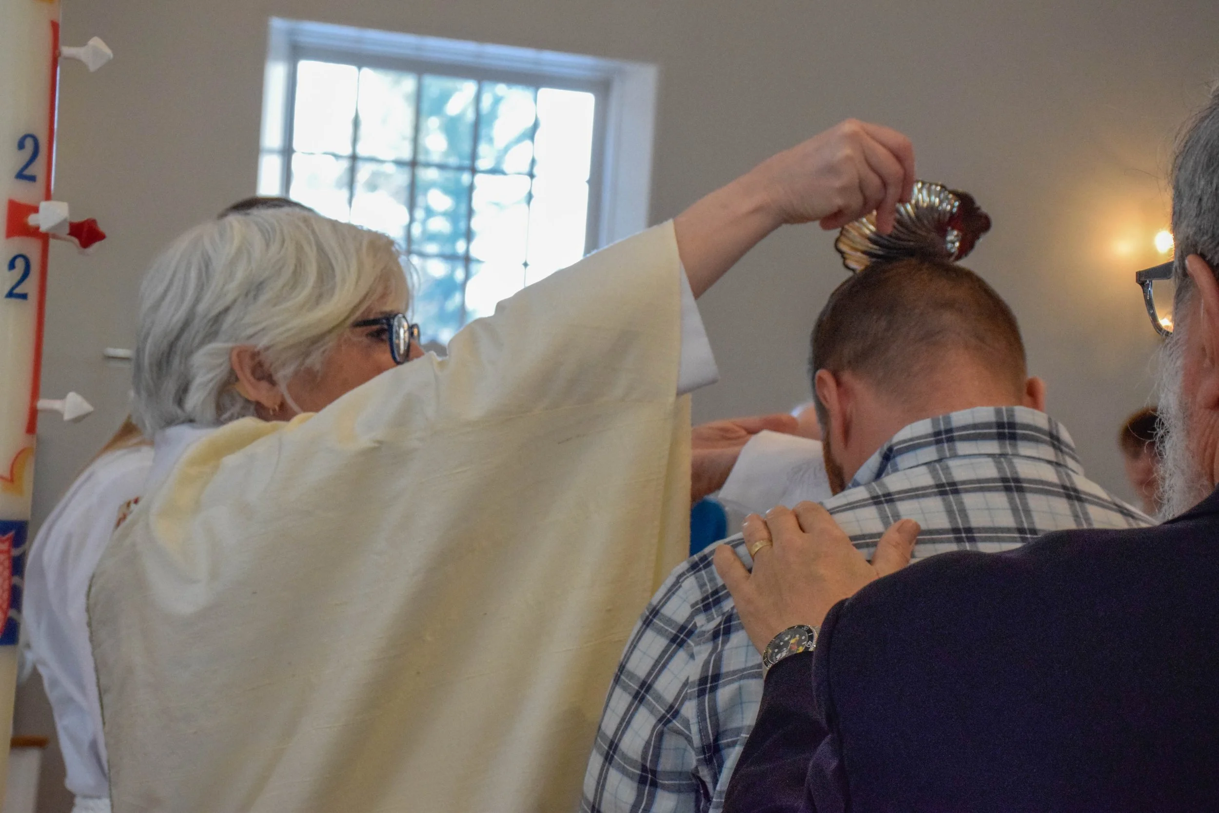 A woman with short gray hair, glasses, and a medical mask holding a seashell on a man's head during a ceremony or event in a room with a window and a clock on the wall.