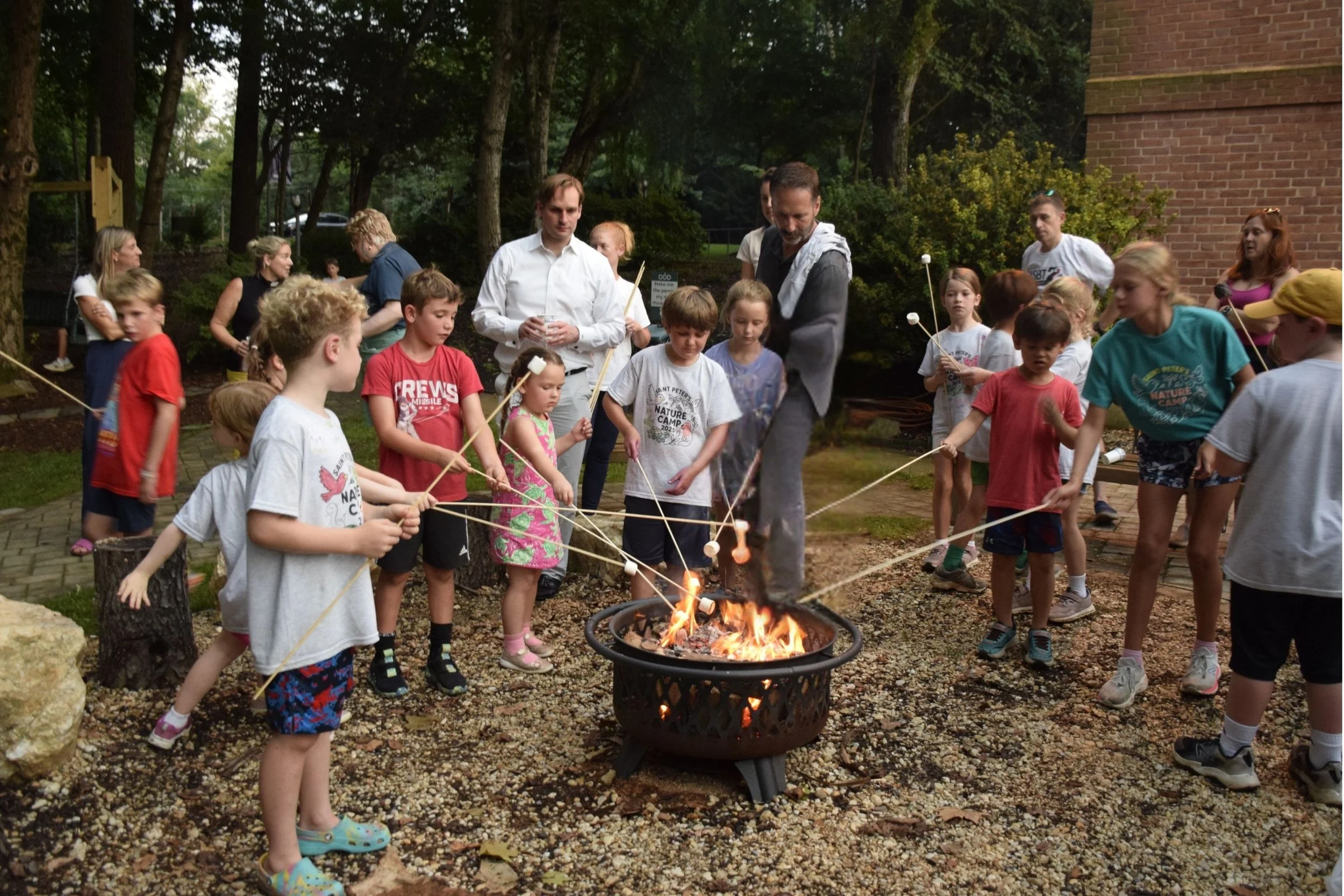 Group of children and adults gathered around a fire pit outdoors, roasting marshmallows on sticks during dusk or evening.
