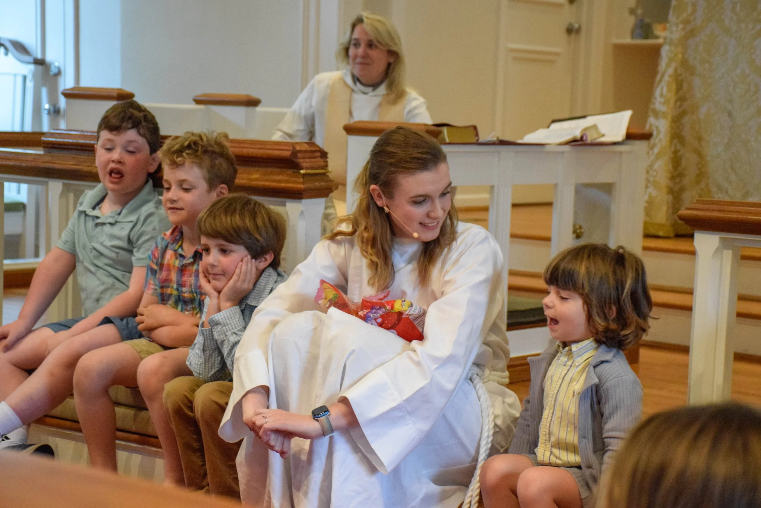 A woman in white robes talking to a young boy in a church setting, with children sitting on a bench nearby