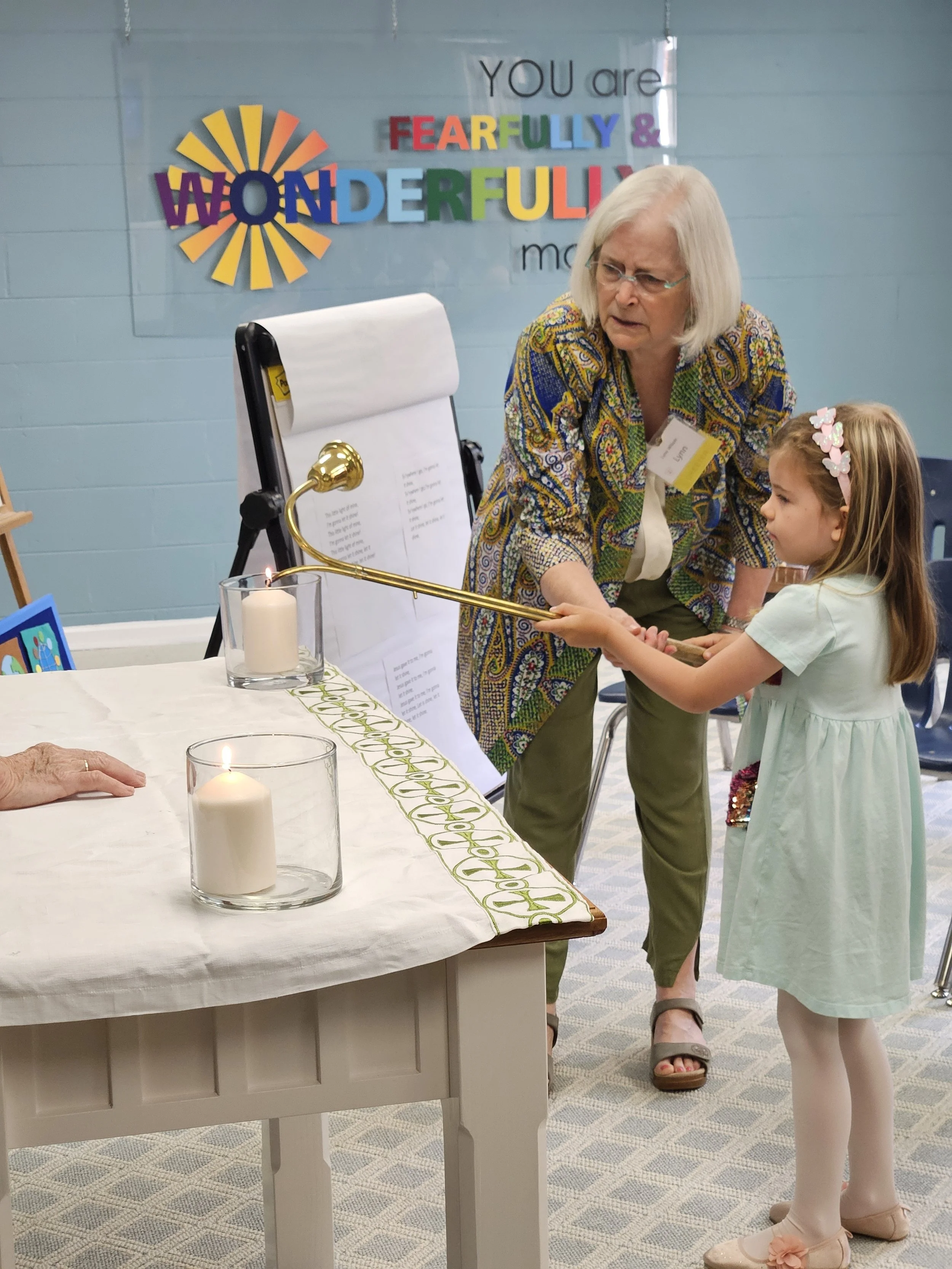 An elderly woman helping a young girl light a candle with a long candlestick to a candle on a table. The table has two lit candles and a decorative cloth. A colorful sign in the background says 'You are wonderfully full of wonder.'