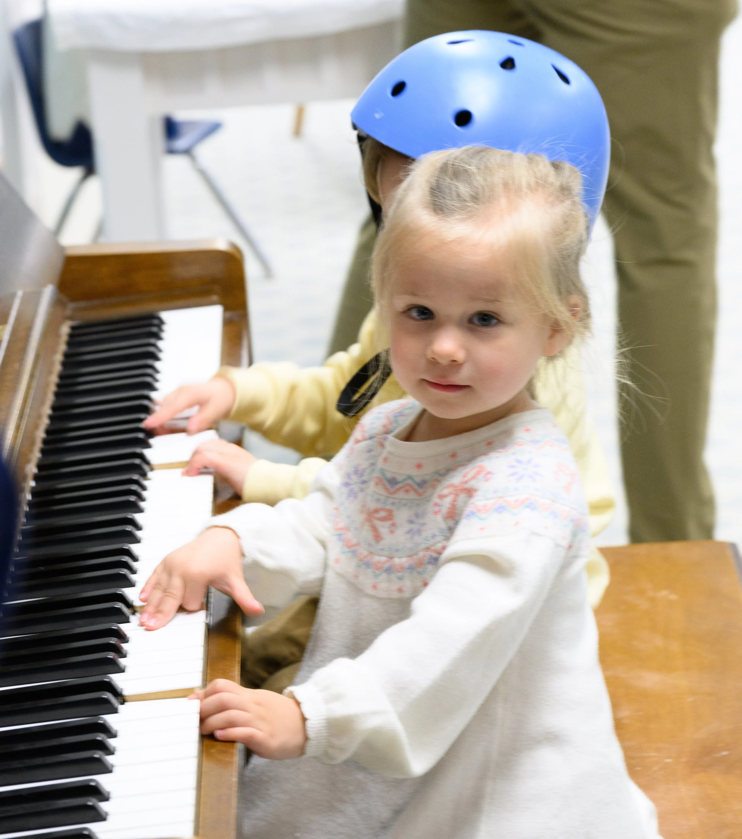 A young girl and a child wearing a blue helmet playing the piano together in a music room.