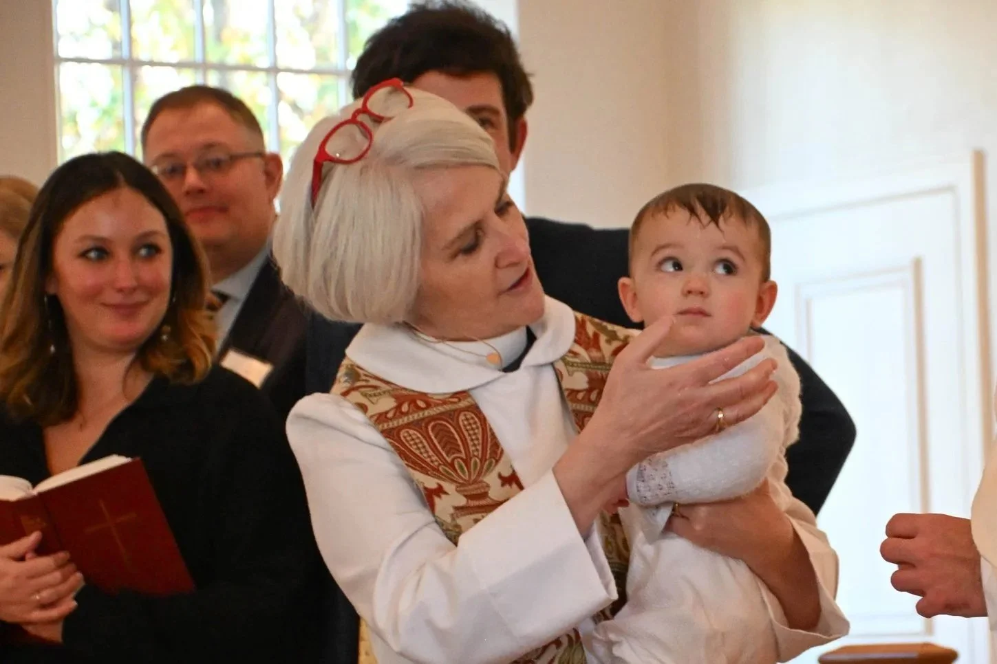 An elderly woman holding a young boy during a religious ceremony or event with several people in the background.