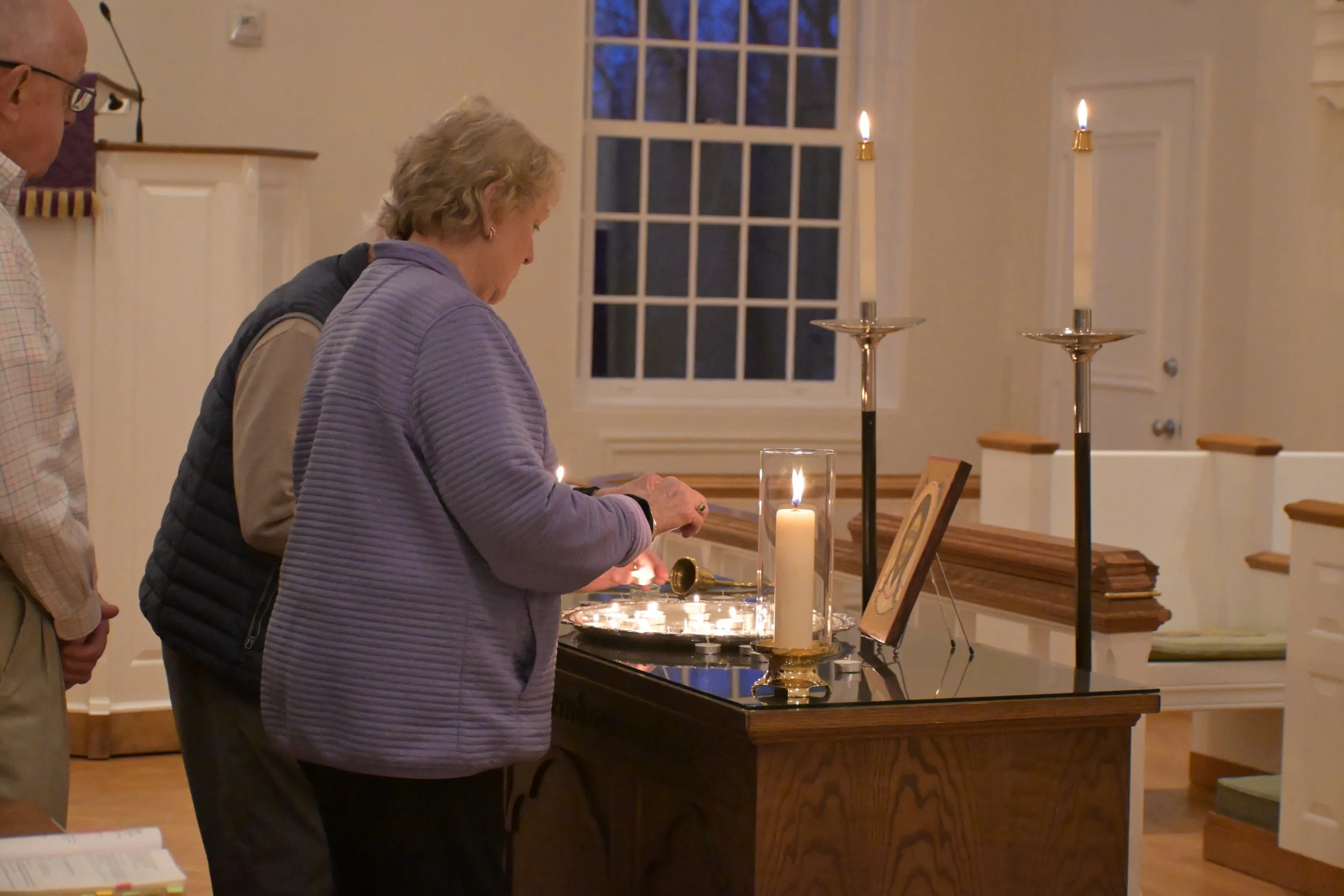 Two women and a man lighting candles during a prayer or memorial service in a well-lit room with a large window and candles on a table.