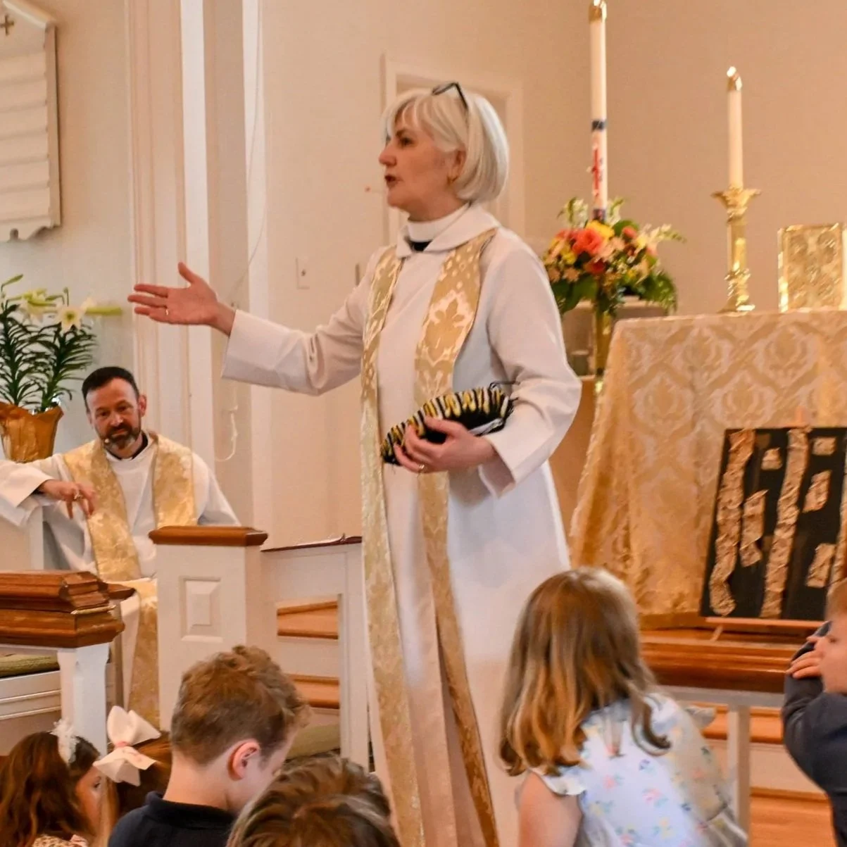 A woman dressed as a priest leading a religious service in a church, with children sitting on the floor listening.