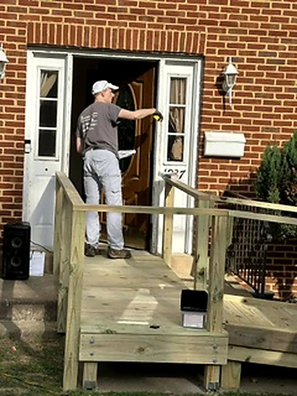 A man installing or repairing the front door of a brick house, standing on a wooden ramp, with tools in his hand and a paper bag in his pocket, outside a house with a lantern and a mailbox.