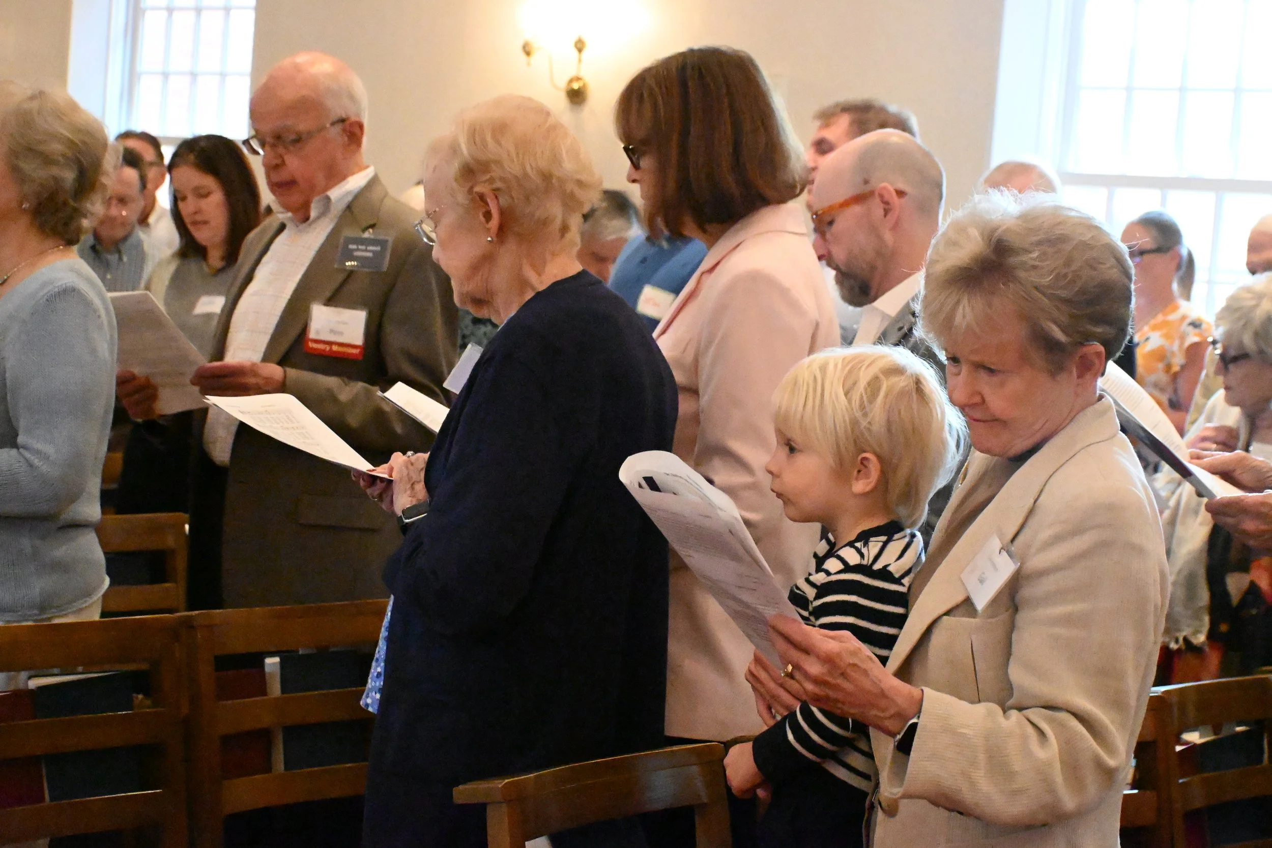 People standing in rows during an indoor event, holding papers and praying or singing with heads bowed.