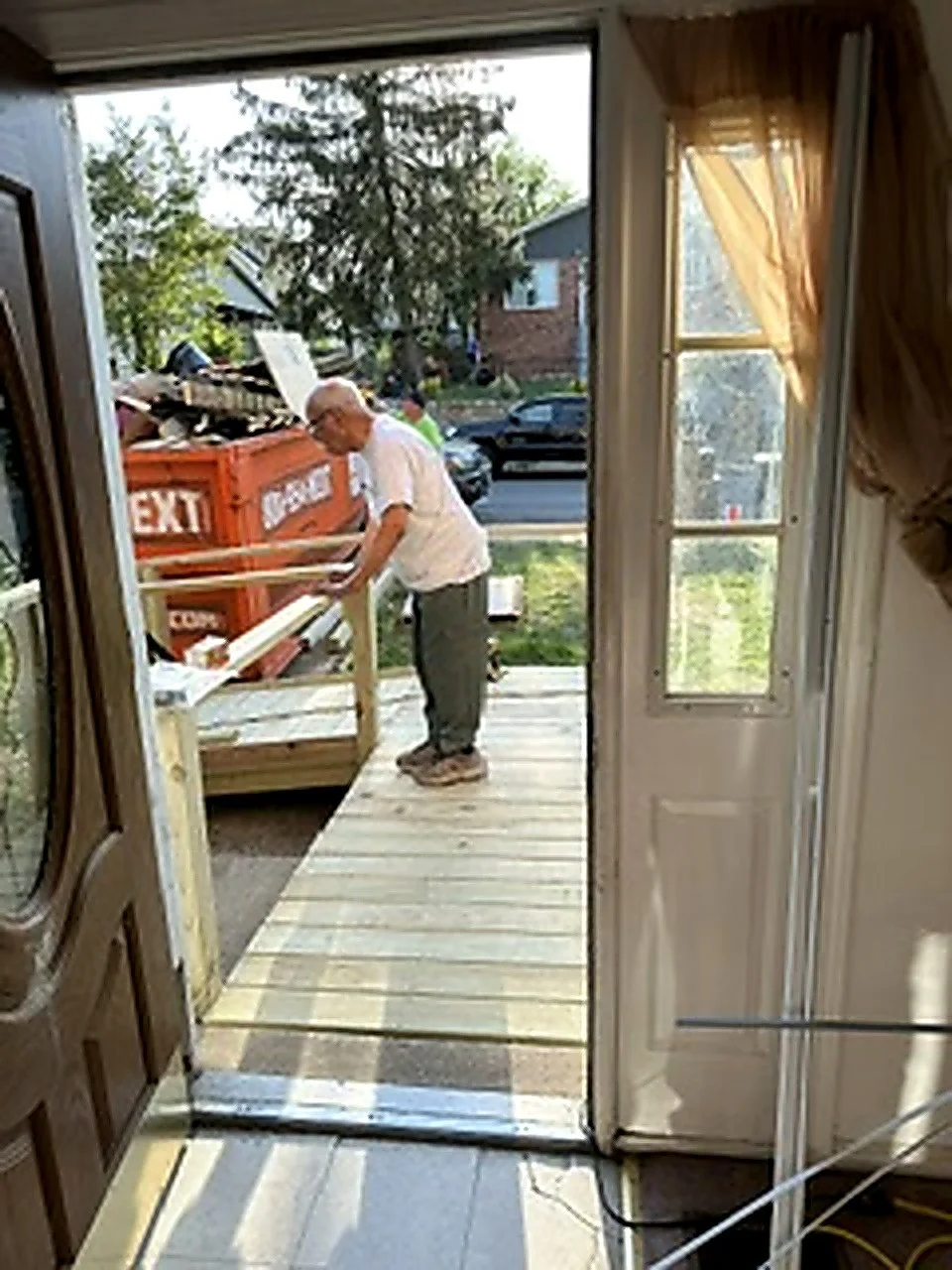 A man is standing outside a house near a door, working on a wooden frame or railing, with a dumpster and trees visible in the background.