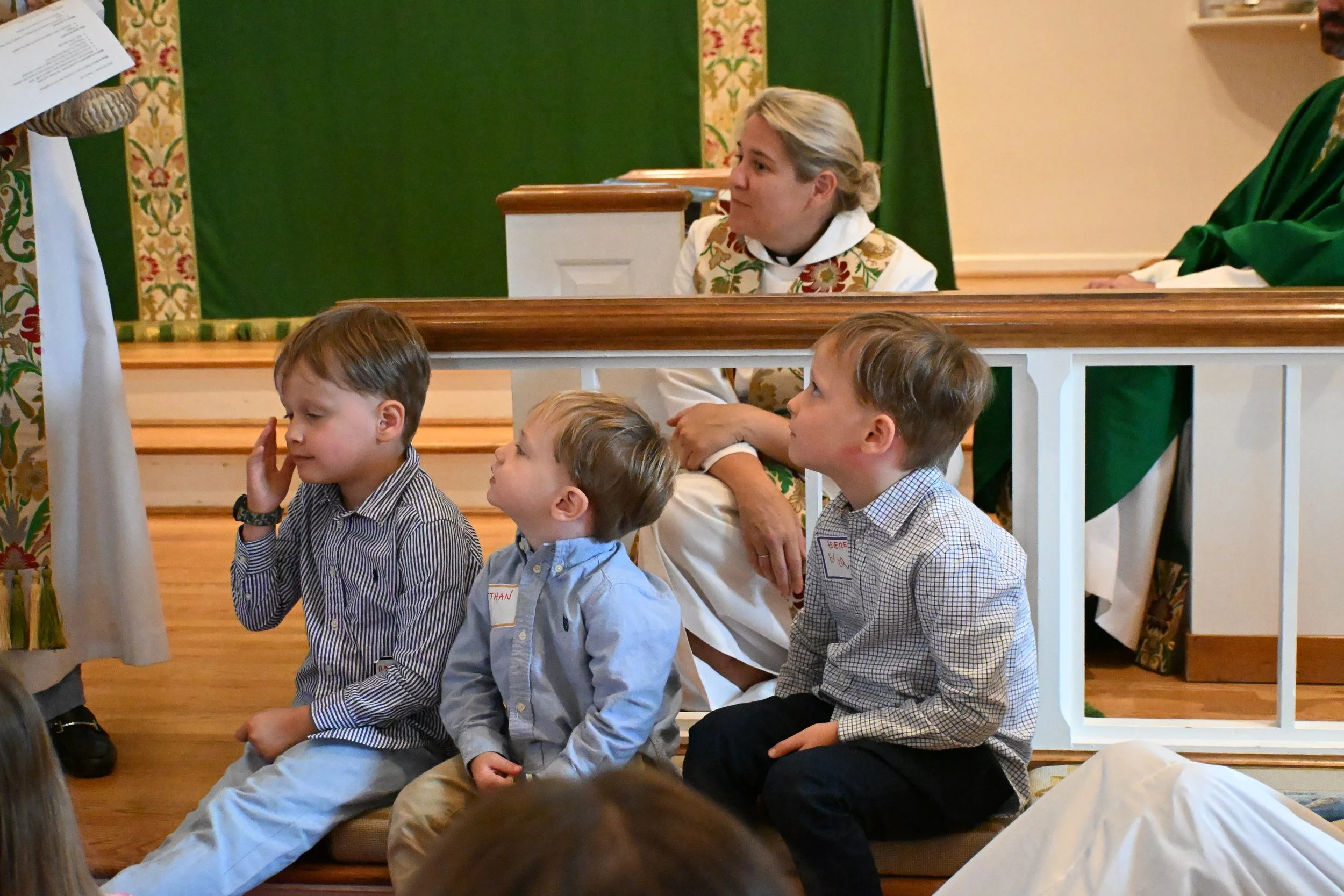 Children sitting on the floor during a church service, with a woman in floral vest sitting behind them, in a church with decorated curtains and floral wallpaper.