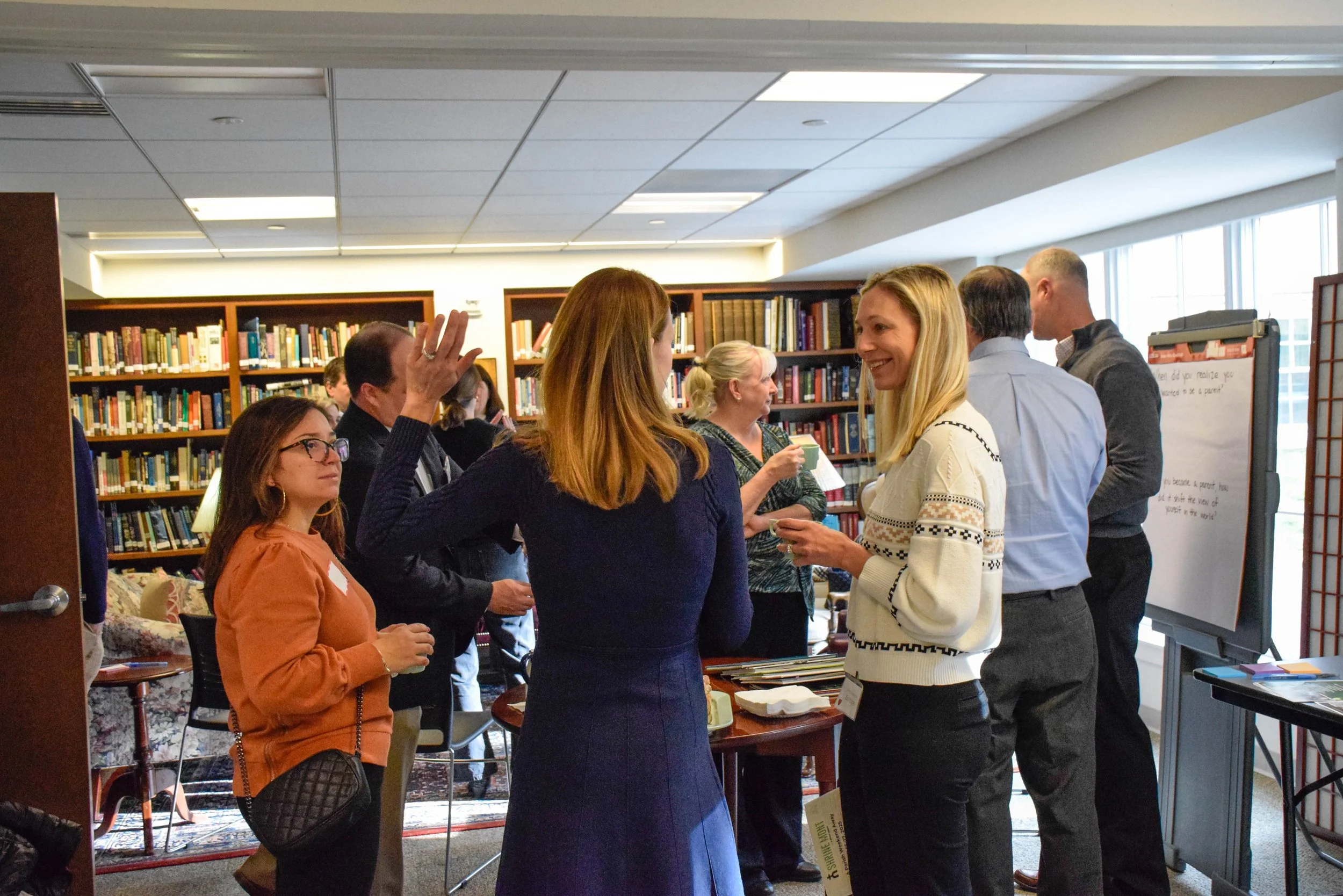 Group of people socializing at a networking event in a library or bookstore, with bookshelves in the background and a whiteboard on the side.