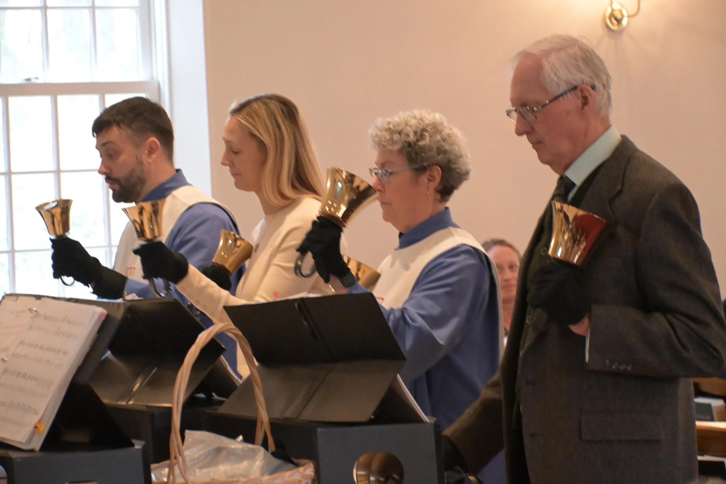 Four people standing in a row holding brass bells during a church service or choir performance, with sheet music and a large window behind them.