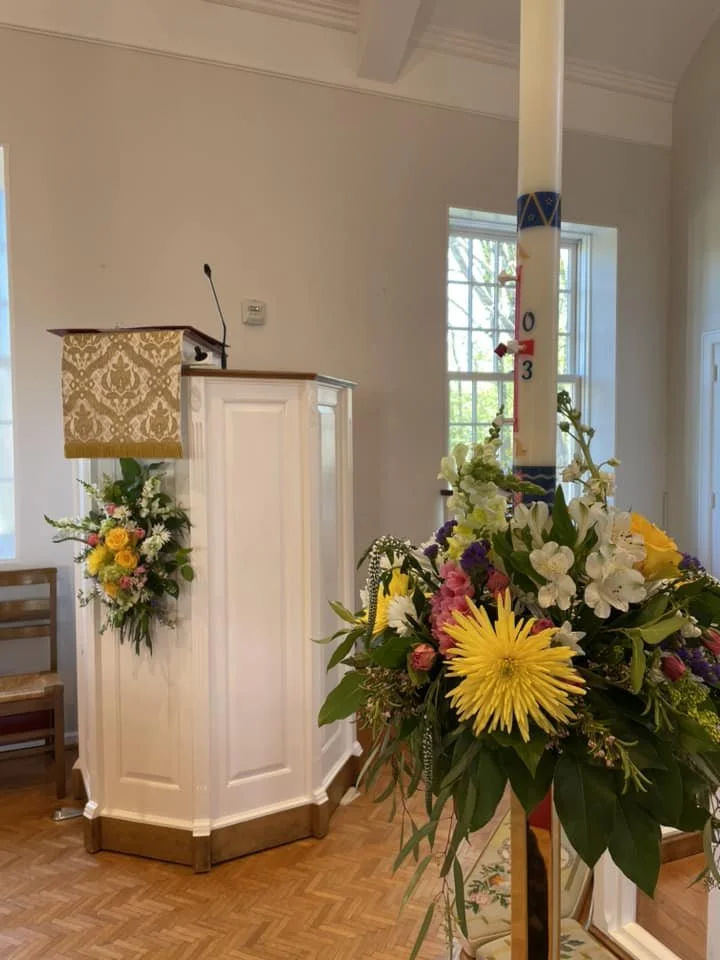 Interior of a church with a wooden pulpit decorated with a floral arrangement, a white candle on a stand with flowers, and a window in the background.