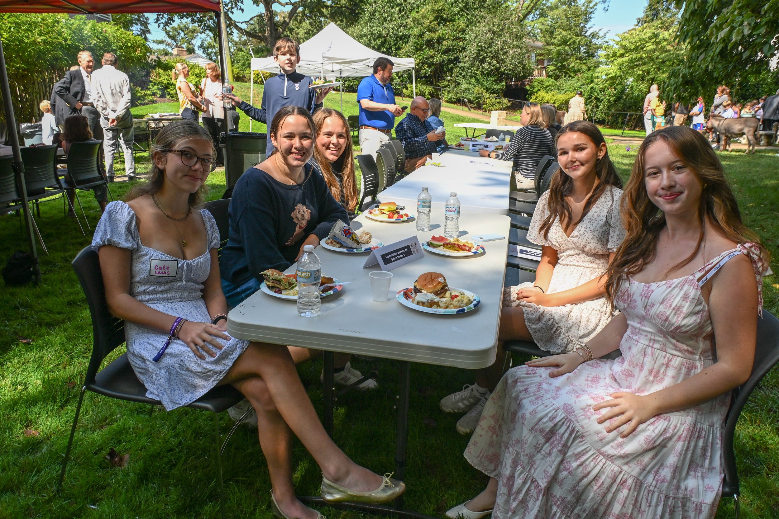 A group of young women sitting at a table outdoors during a sunny day, with more people in the background at what appears to be a celebration or gathering in a garden.