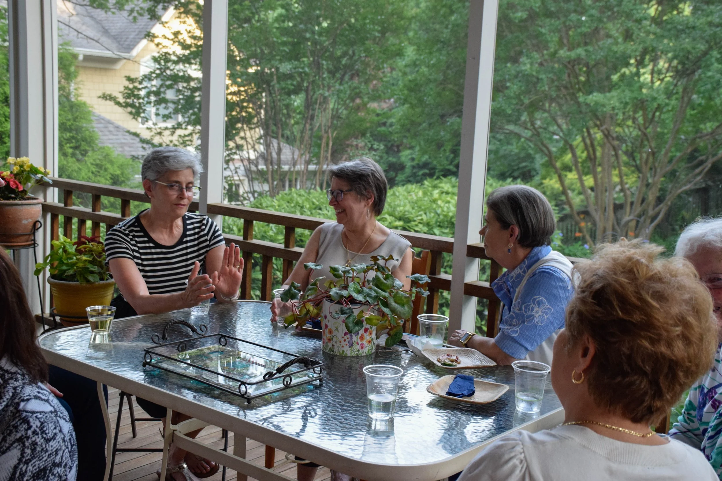 A group of older women sitting around a table on a screened porch, engaging in conversation and laughter, with outdoor greenery visible in the background.