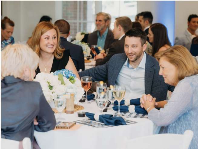 People dining at a banquet table, smiling and conversing in a bright, modern event space.