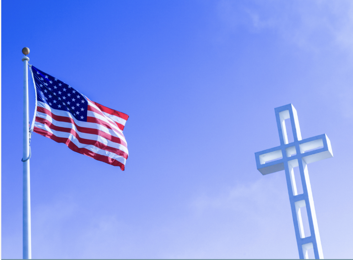 American flag flying next to a white illuminated cross against a blue sky.