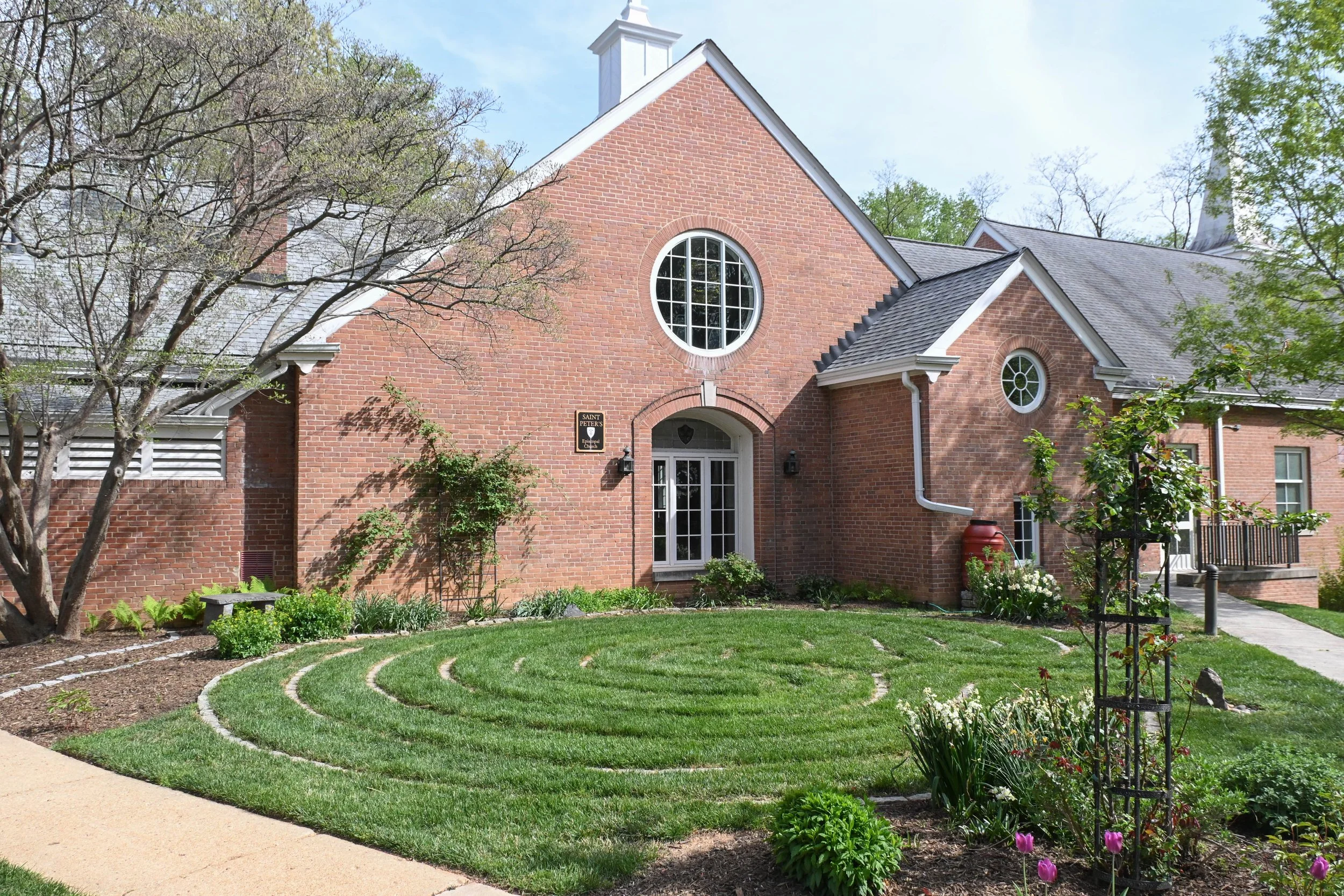 A red brick church with a gabled roof, circular windows, a white door, and a manicured lawn with a spiral pattern. There are trees and flowering plants in the garden.