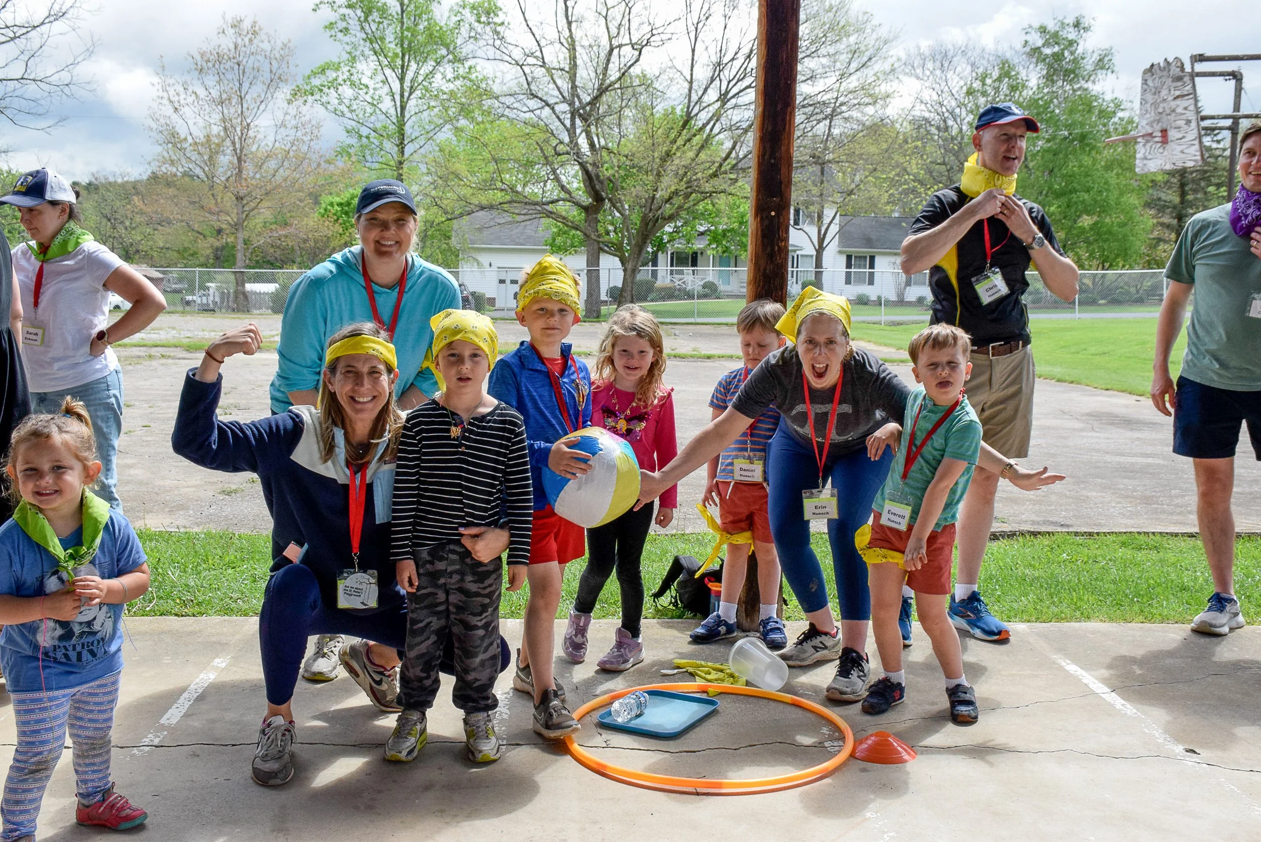 Group of children and adults outdoors for a team activity, some children wearing yellow bandanas, with a sports ball, hula hoop, and cones on the ground.