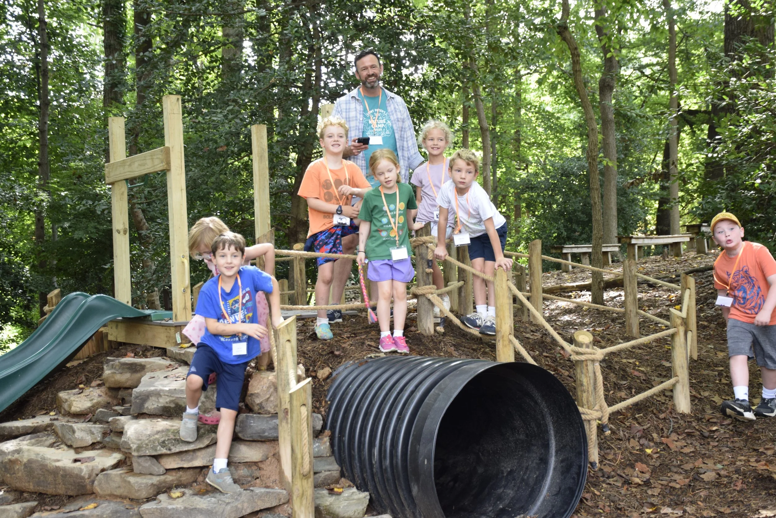 Group of children and a man outdoors on a wooden playground structure, surrounded by trees, with a large black tunnel and a slide.