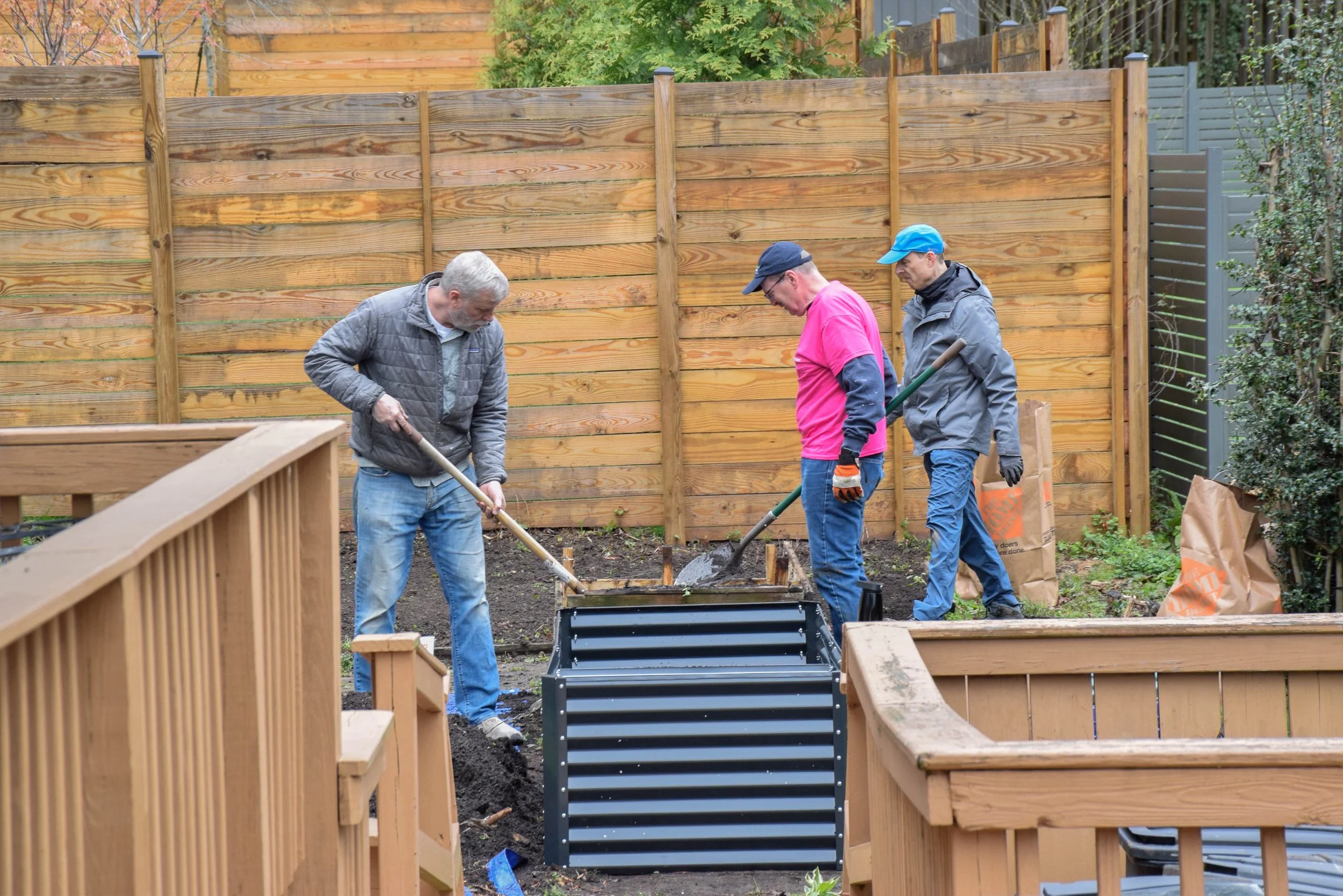Three men working on a gardening or construction project in a backyard, with a wooden fence in the background. They are using shovels to till or dig in the soil.
