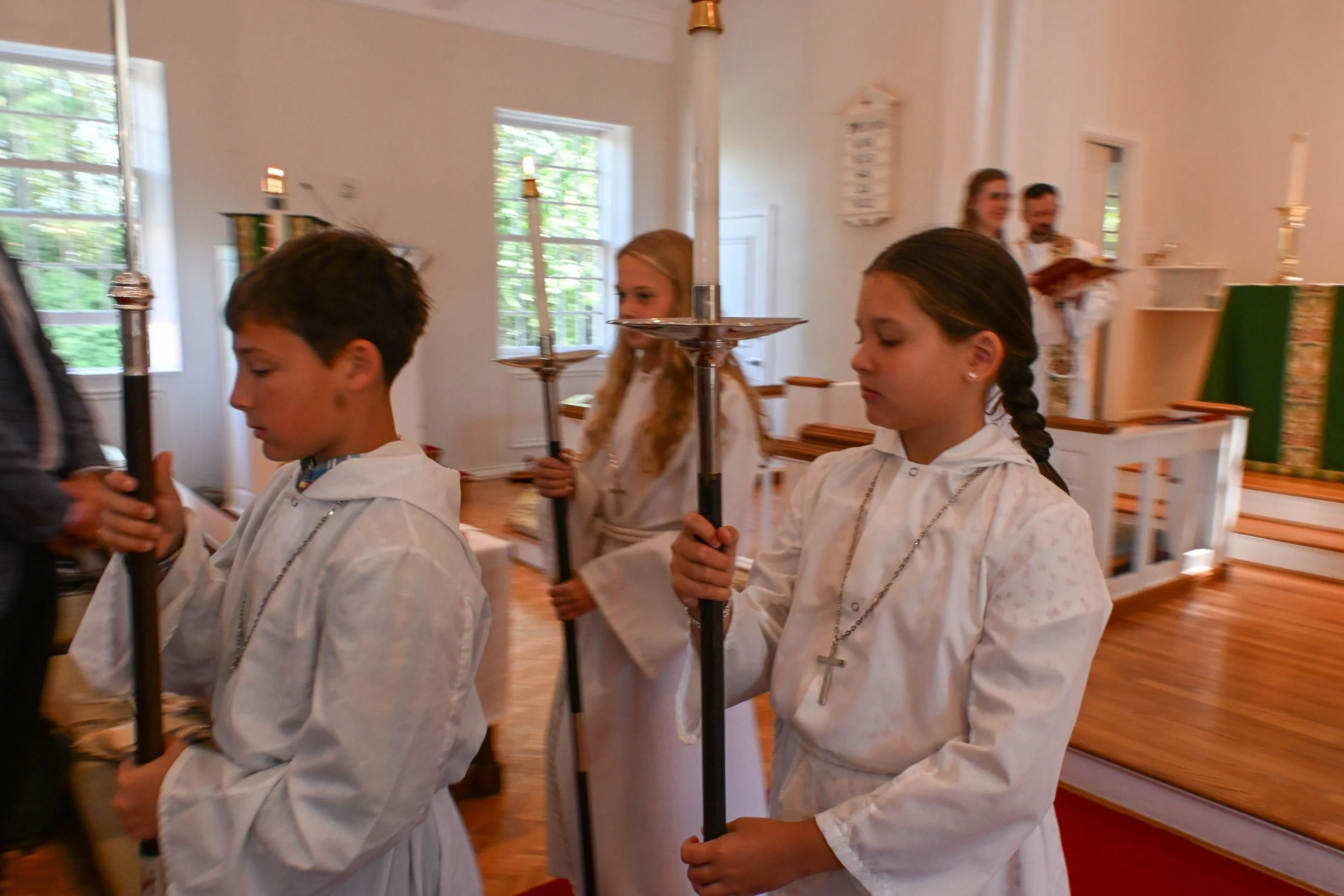 Children participating in a church ceremony, holding candles, inside a church with large windows and wooden pews.