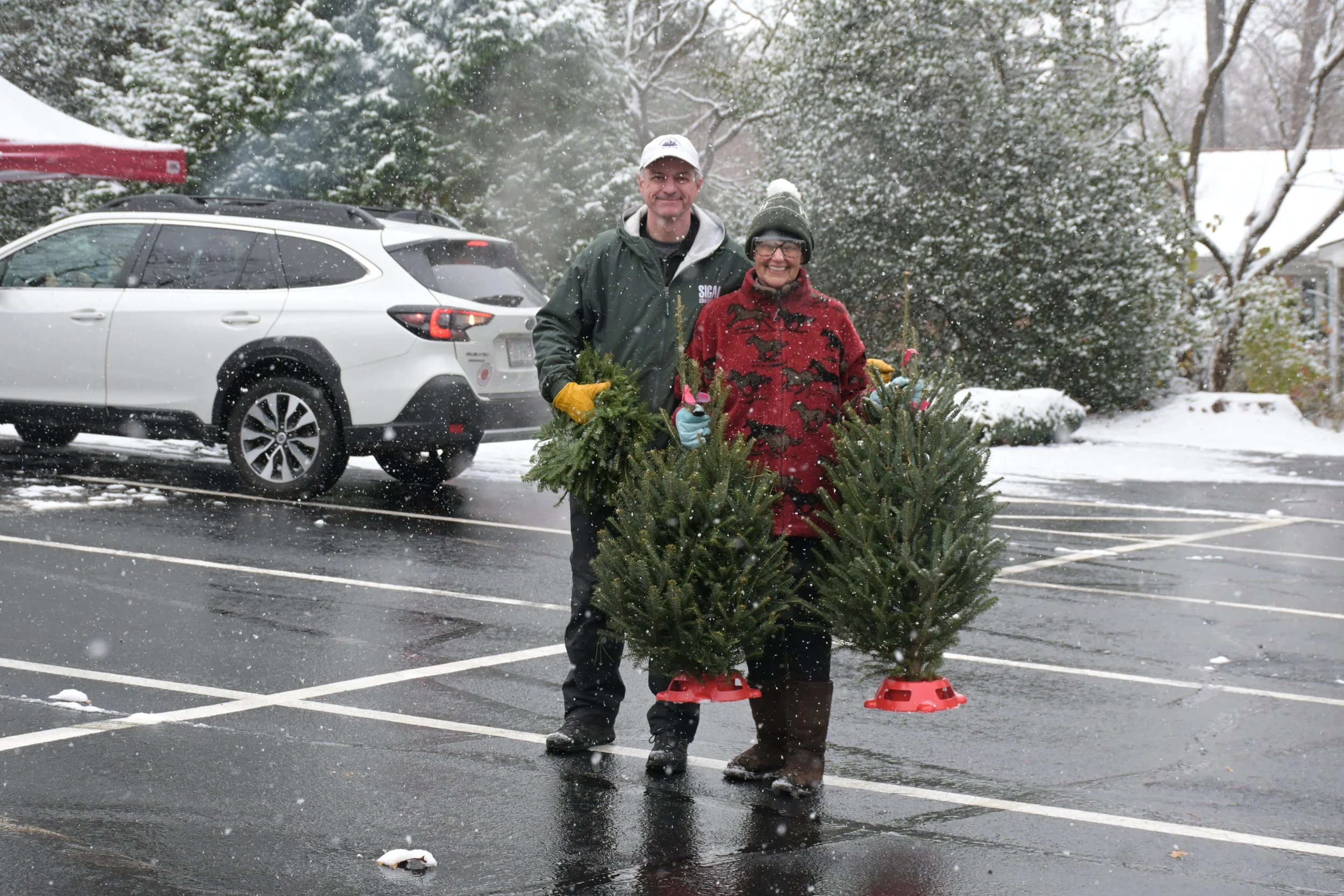Two people standing outside in a parking lot during snowfall, holding small Christmas trees with red tree stands, smiling at the camera.