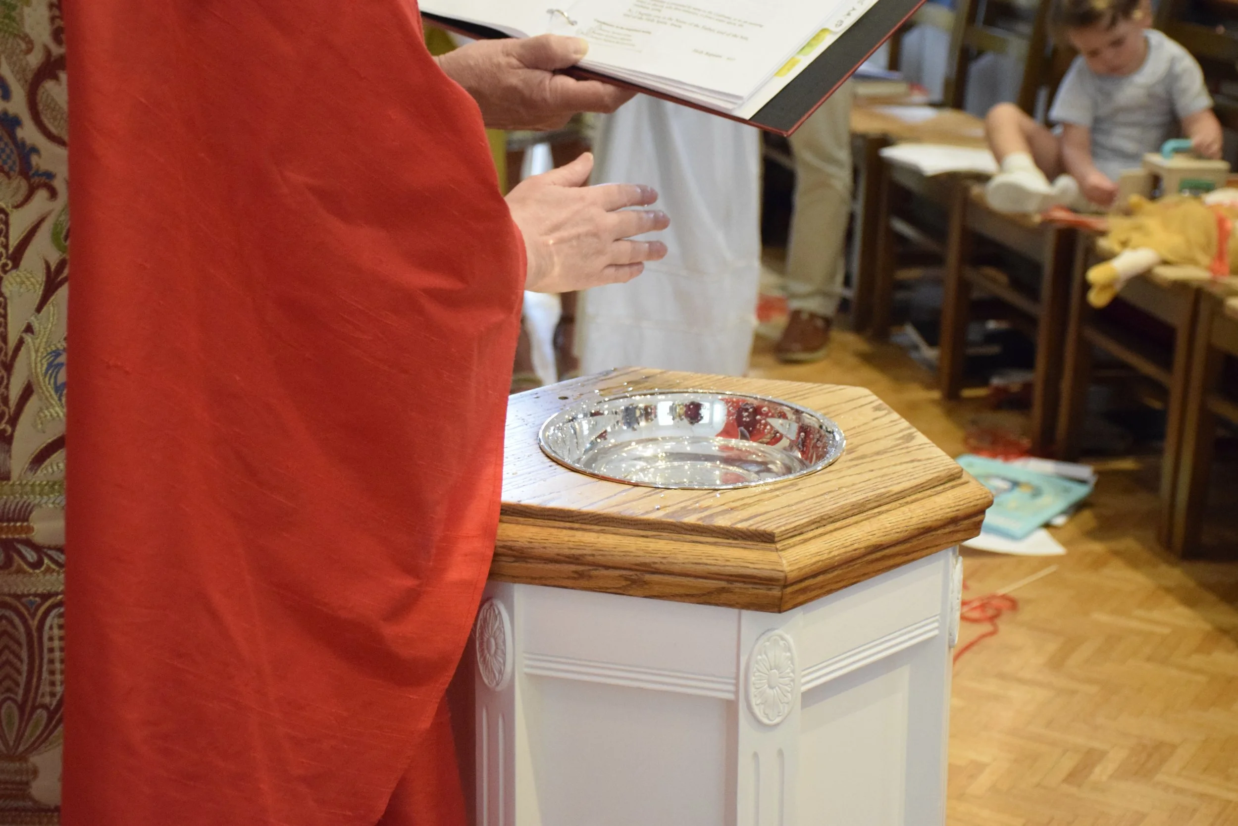Person in a red shirt standing at a wooden table with a silver bowl, holding a binder or book, with children and tables in the background.
