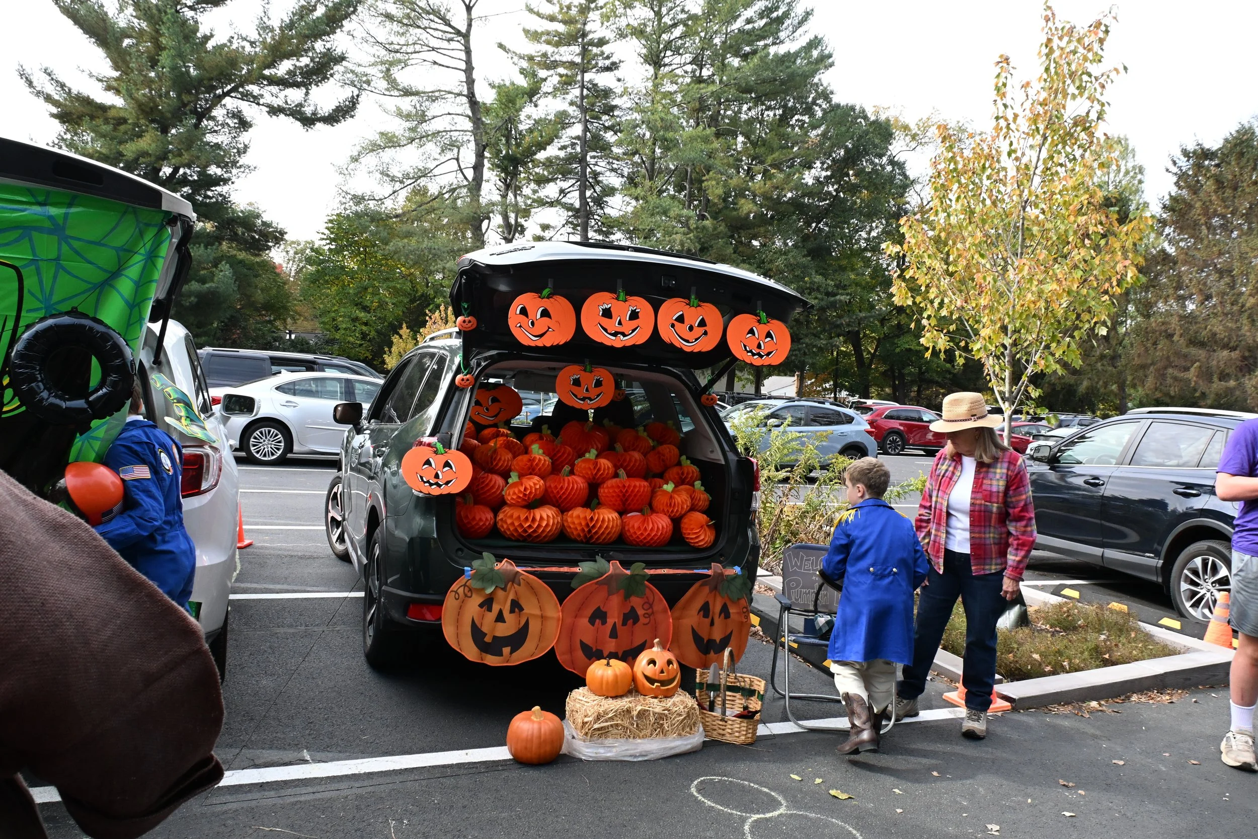 A parking lot decorated for Halloween with a black hatchback car filled with orange pumpkins. Halloween-themed decorations, including paper lanterns and hanging pumpkins, adorn the vehicle. A woman and two children dressed in Halloween costumes are nearby, with the children collecting or purchasing pumpkins.