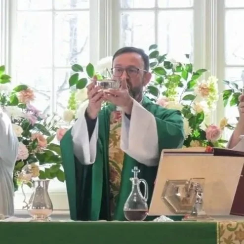 A man dressed in religious vestments holding a chalice during a church ceremony in a bright room with large windows and floral arrangements.