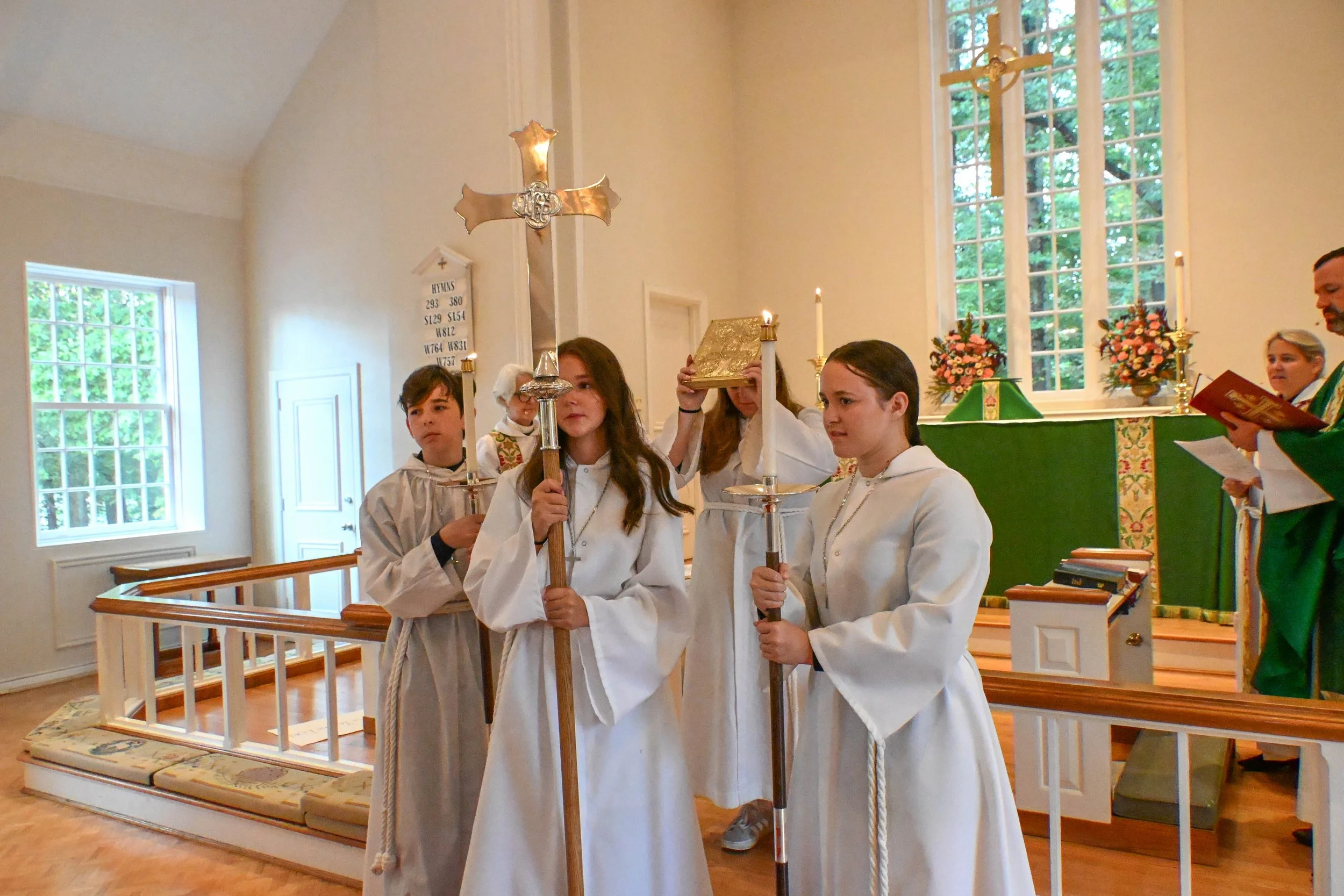 A group of children and a woman participating in a religious ceremony inside a church. They are dressed in white robes, holding candles and a Bible. The church has large windows, floral arrangements, and an altar with candles and religious decorations.