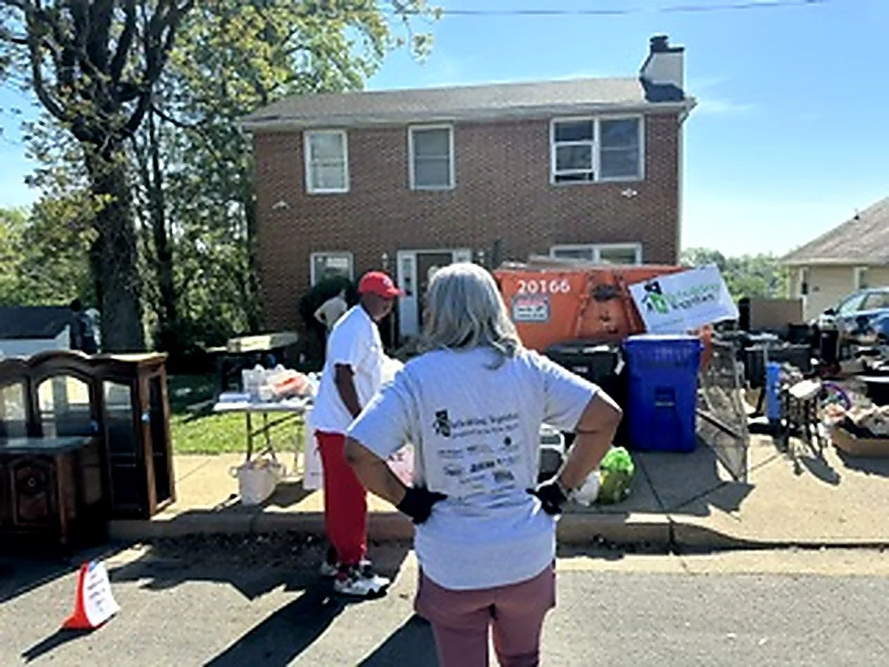 People participating in a community cleanup event outside a house, with supplies, a trash bin, and furniture on the sidewalk on a sunny day.