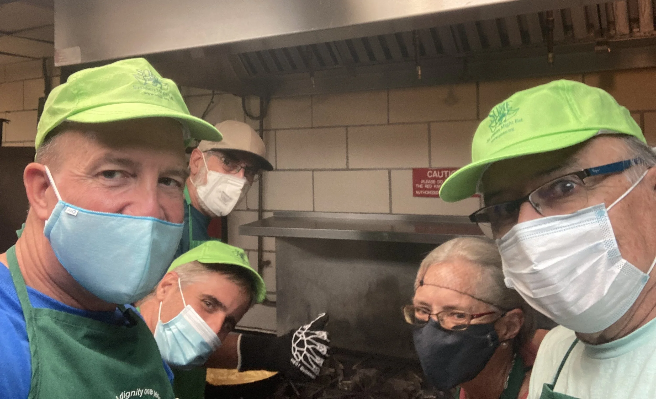 Group of five people wearing masks and green hats, working in a kitchen. They appear to be volunteering or working together, with one person giving a thumbs up.