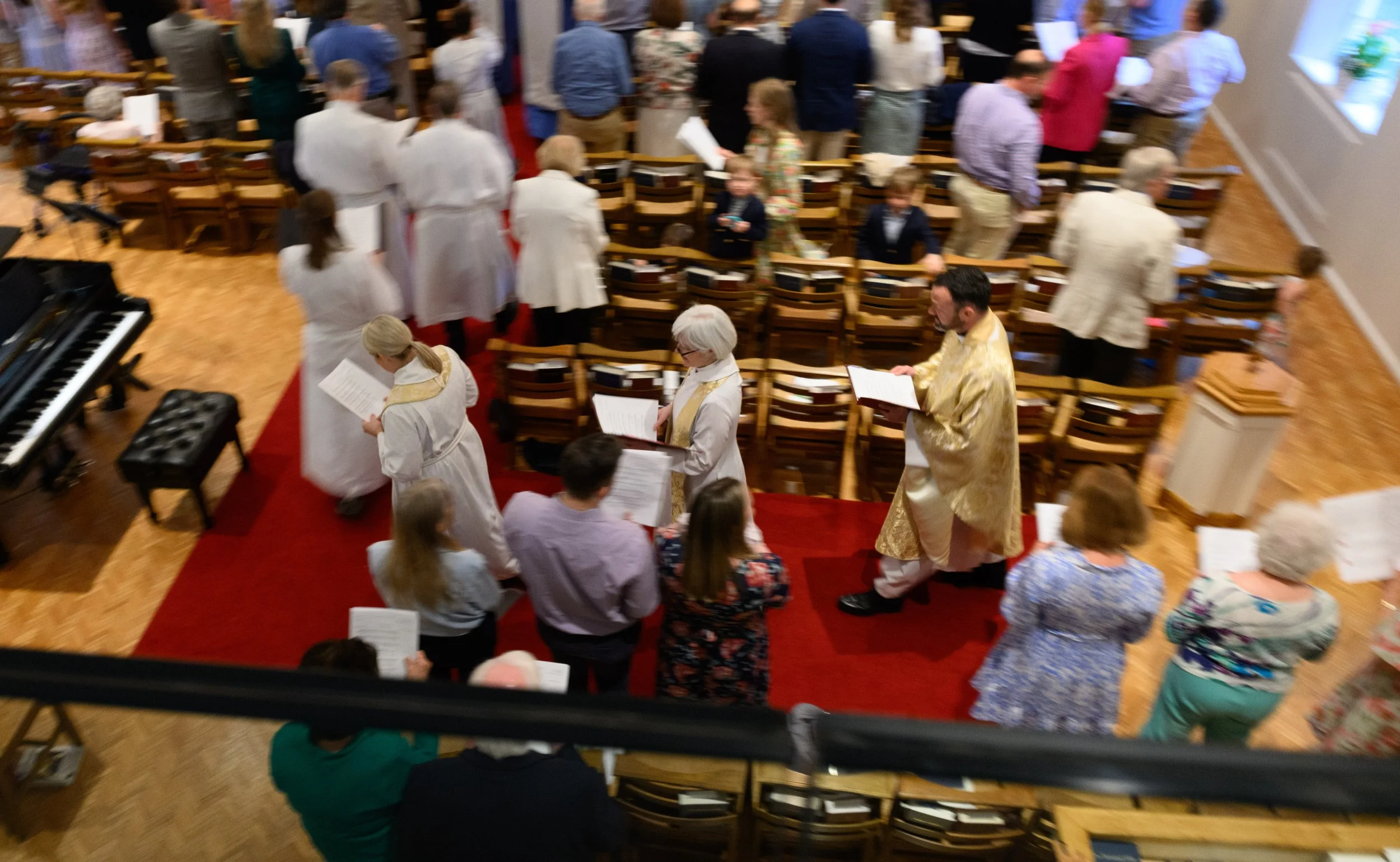People attending a church service or choir practice, some standing and reading from music sheets, others sitting in pews, with a piano visible on the left side of the image, all in a brightly lit, spacious room.