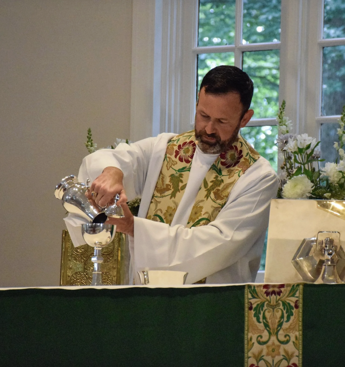 A man dressed in religious robes, with a patterned stole, pouring water from a silver pitcher into a silver bowl in front of him during a religious ceremony inside a room with a window and flowers.