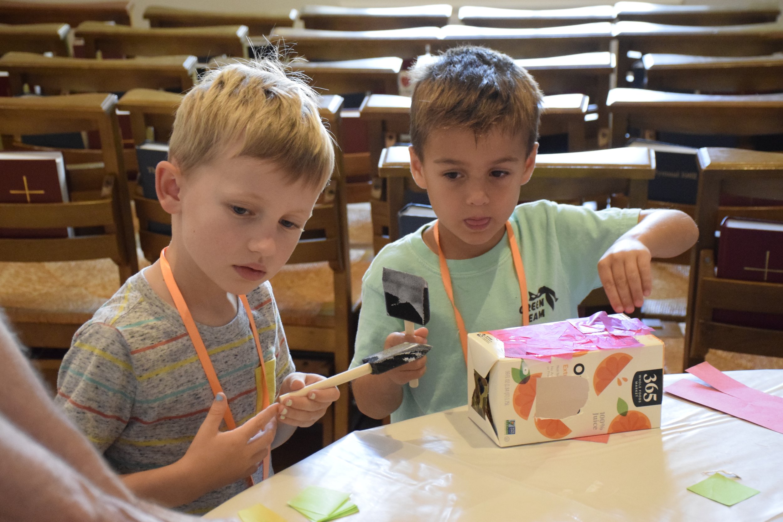 Two young boys working on craft projects at a table in a room with wooden chairs, one boy holding a paintbrush and the other holding a paint palette, with a carton of juice on the table.