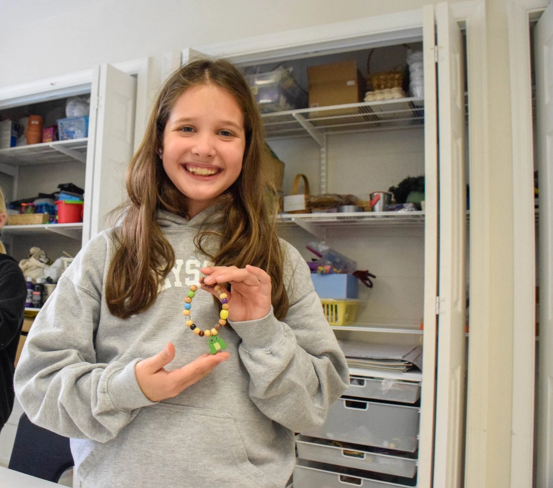 A smiling girl in a gray hoodie holding a colorful beaded bracelet with a green turtle charm in front of open storage shelves.