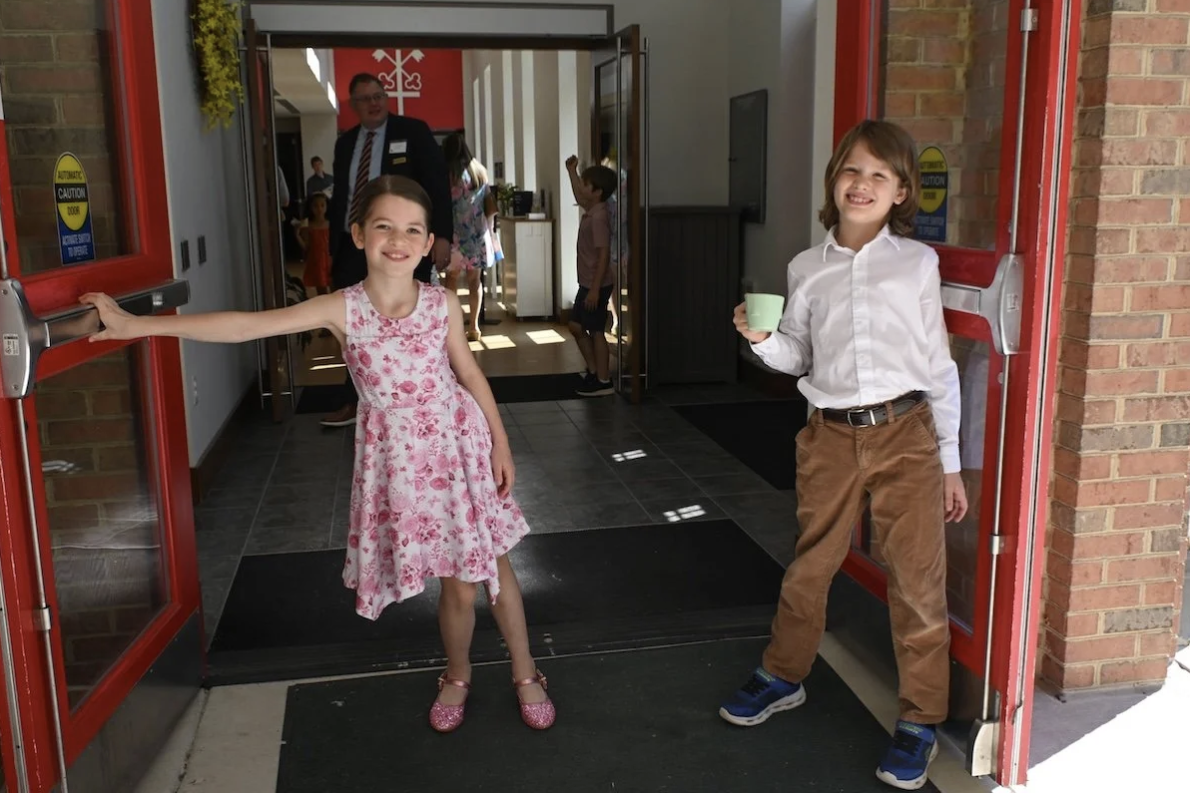 Two children, a girl in a pink floral dress and a boy in a white shirt and brown pants, standing at the entrance of a building, smiling.