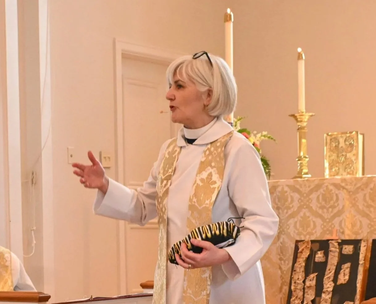 An elderly woman with short white hair, wearing glasses on her head, a white garment with a gold embroidered vest, is speaking inside a church. She holds a small purse with a black and yellow tiger stripe pattern. Behind her are lit candles on gold candleholders, floral arrangements, and religious icons.