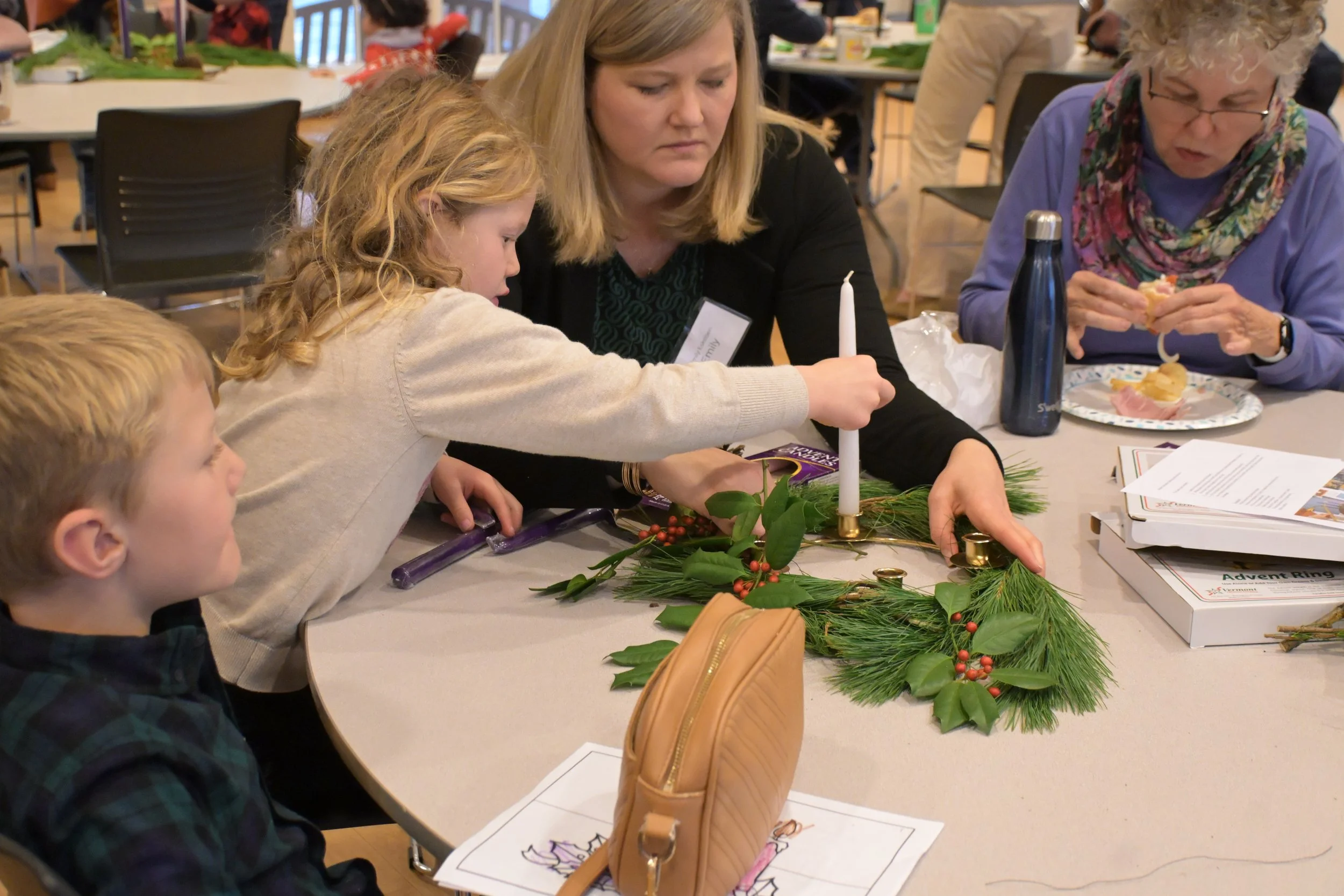 People making holiday wreaths at a craft table. A woman helps a young girl as they work on decorating a wreath with greenery and red berries, with other individuals seated nearby.