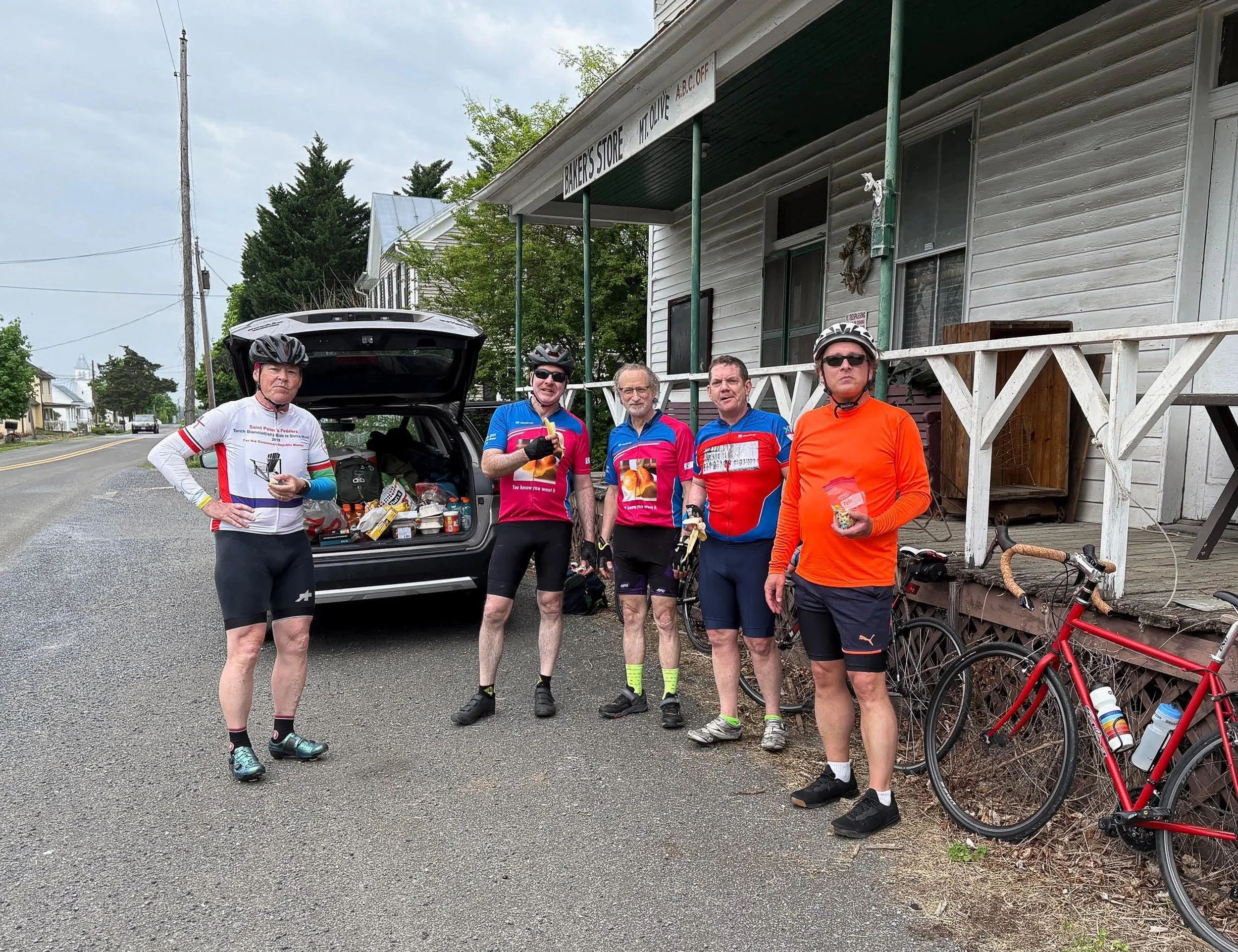 Five cyclists in colorful gear standing outside a house with an open car trunk filled with supplies, two red bicycles leaning against a porch railing, on a cloudy day in a small town.