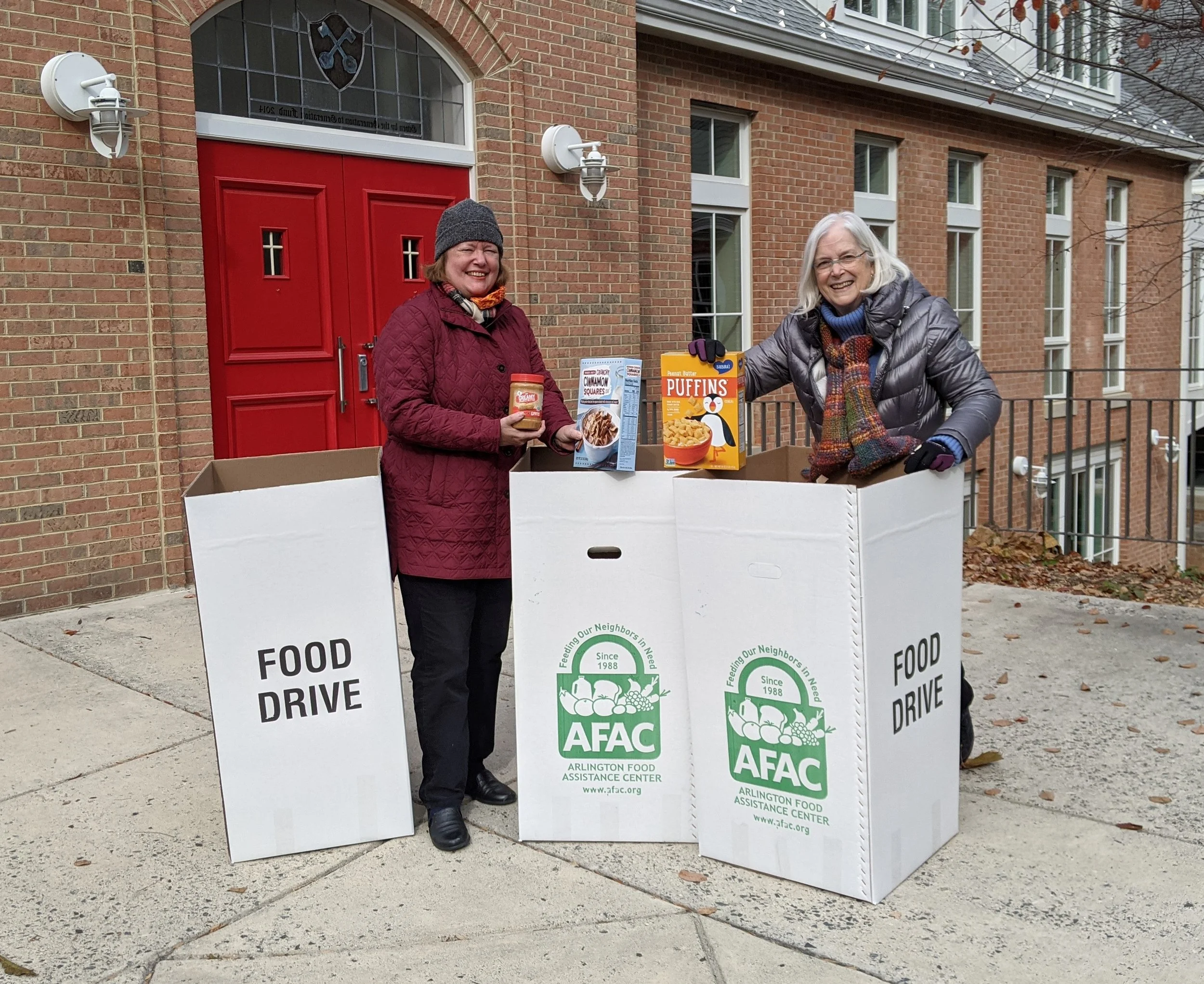 Two women standing behind a large white food drive donation box with green and black text and logo, smiling at the camera, outside a brick building with a red door and multiple windows, during fall with some leaves on the ground and tree branches overhead.