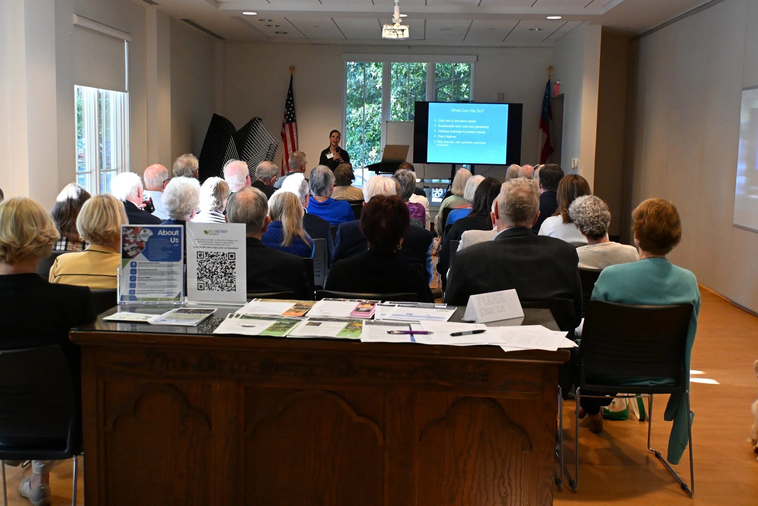 A woman is giving a presentation to an audience in a conference room. The speaker is standing next to a screen displaying a slide titled 'What Can We Do?' with bullet points. The room is filled with seated attendees, and there are tables with pamphlets, brochures, and QR codes in the foreground.